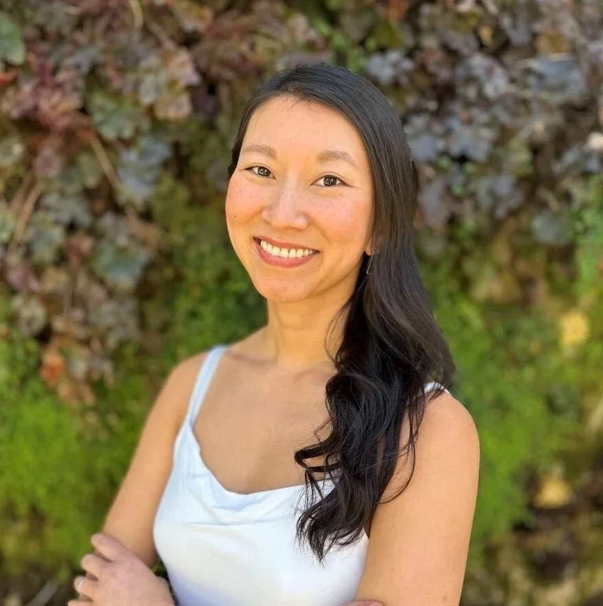 A woman with long dark hair, smiling, wearing a white sleeveless top, standing outdoors in front of a background of plants.