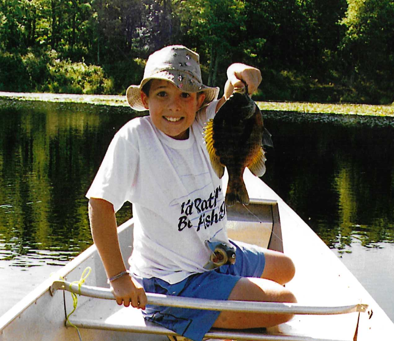A young boy sitting in a boat holding a large fish he caught, on a calm lake with trees in the background.