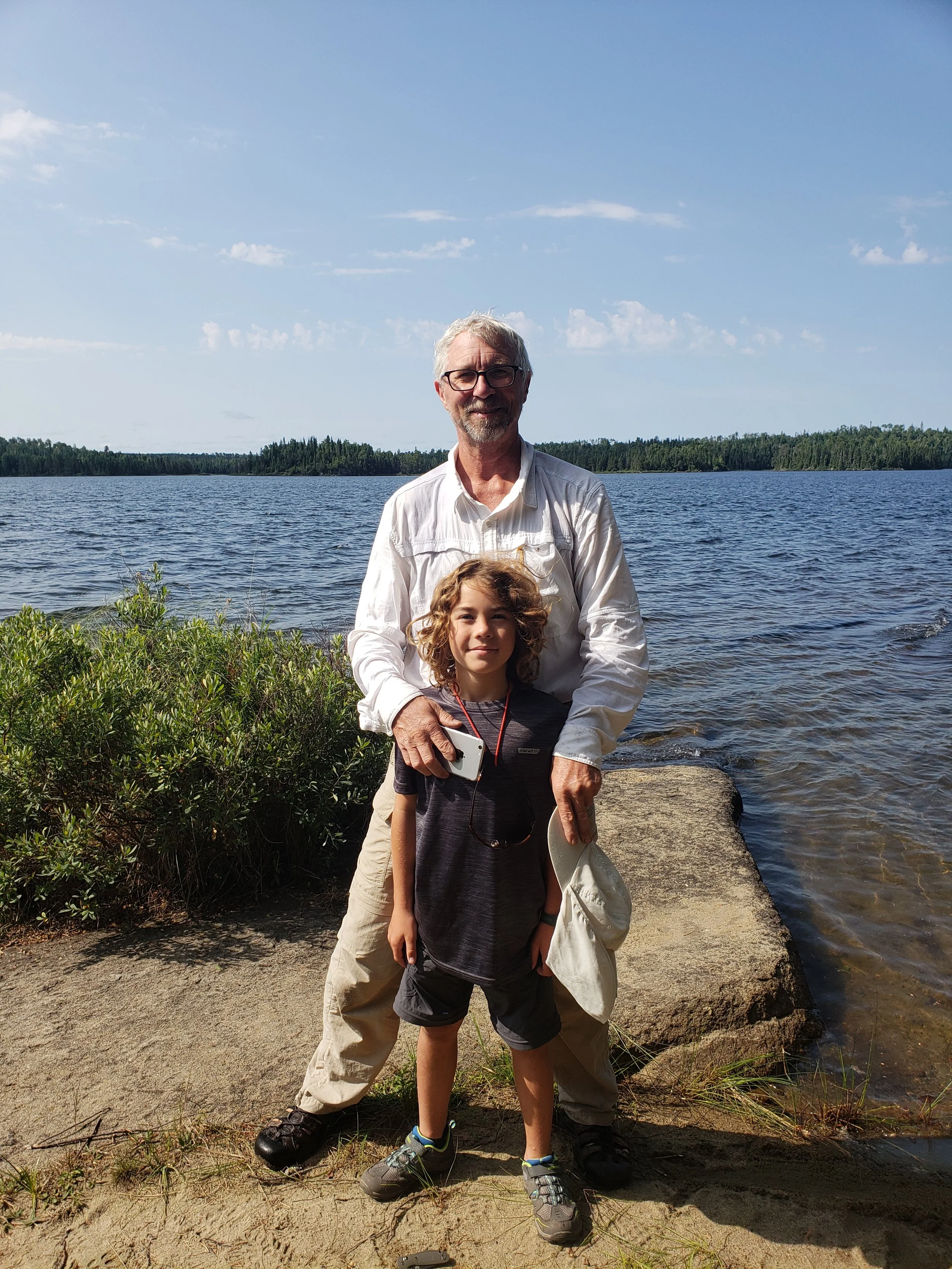 A man and a child standing water's edge with a lake and forest in the background on a sunny day.