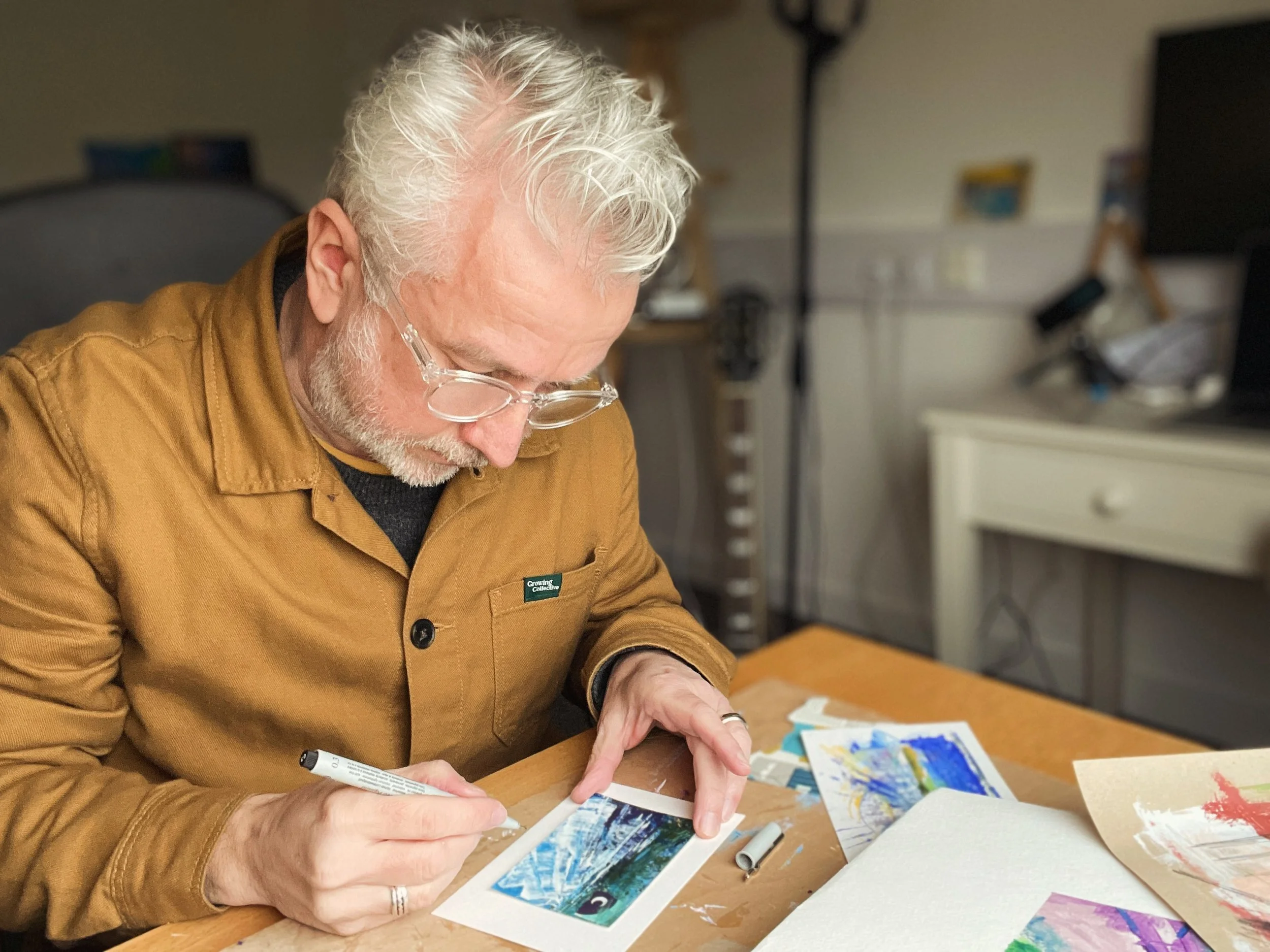 An artist is sitting at a table, engaging in painting or colouring on paper with a marker pen. On the table, there are colorful artworks, and in the background, art supplies and a lamp are visible. The man is wearing a brown jacket.
