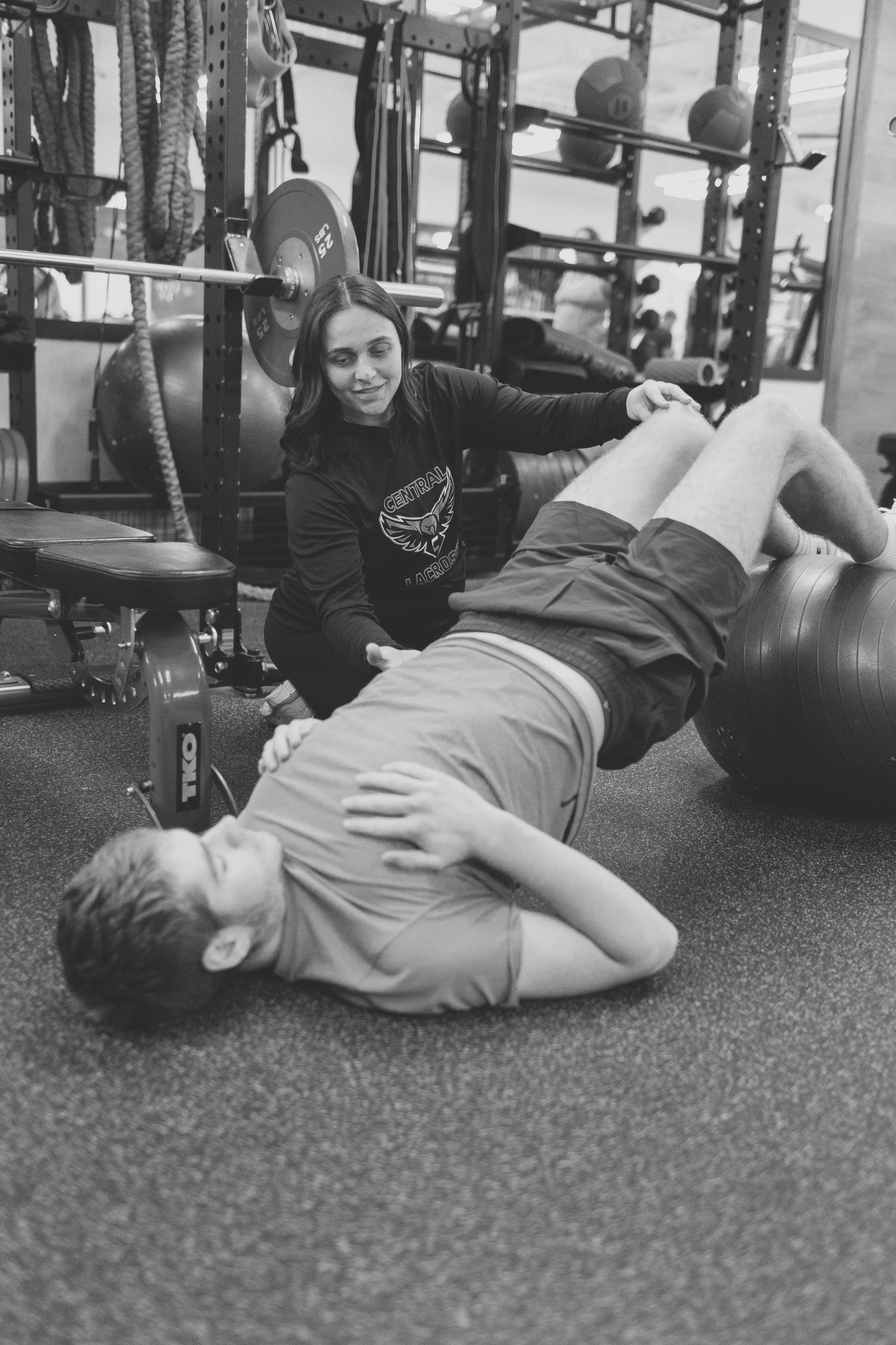 A woman appears to be a trainer or physical therapist, assisting a young man performing an exercise or mobility drill on the floor in a gym. The young man is lying on his side with his head resting on his arm, while the woman is supporting his hip with her hand. The gym background includes weight plates, resistance bands, medicine balls, and other fitness equipment.