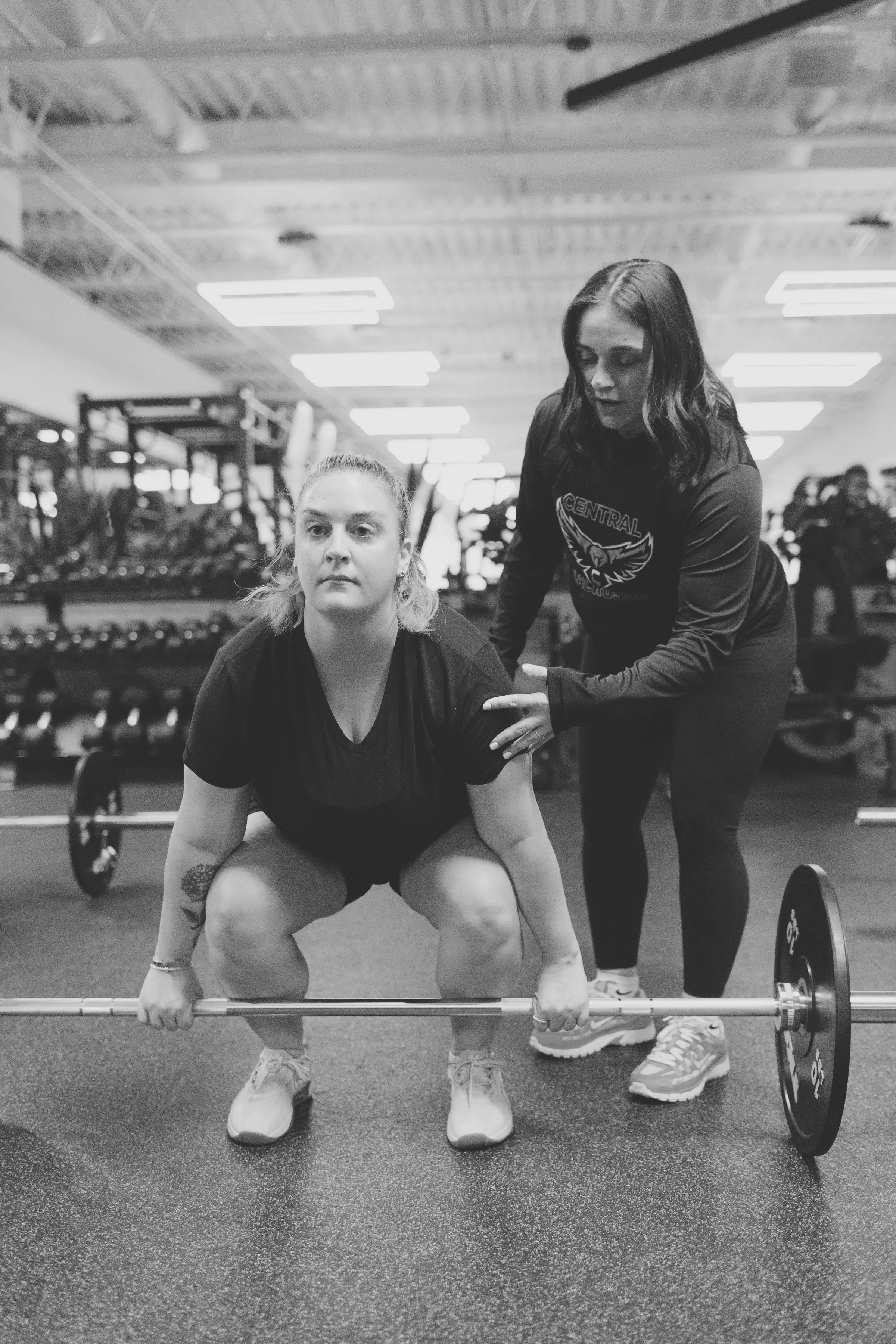 Two women in a gym, one is squatting with a barbell, while the other stands behind her providing support. The woman squatting has her hair tied back, and the woman standing has shoulder-length hair. Gym equipment and other people working out are visible in the background.