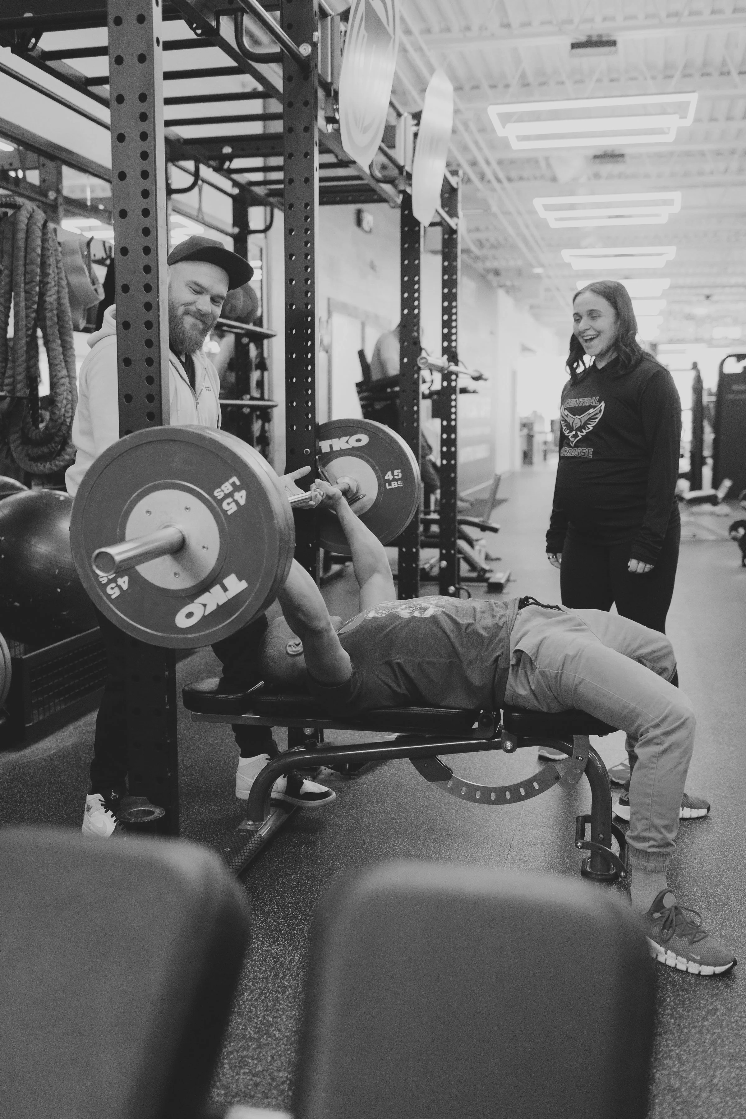 A man is lying on a workout bench performing a bench press with a barbell, while another man stands behind him spotting, and a woman is standing nearby, smiling at the scene, in a gym with exercise equipment in the background.