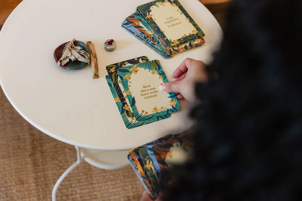 A person holding a deck of cards with a tropical leaf pattern near a table with other cards, a small round decorative object, a stick, and a ring with a red stone.