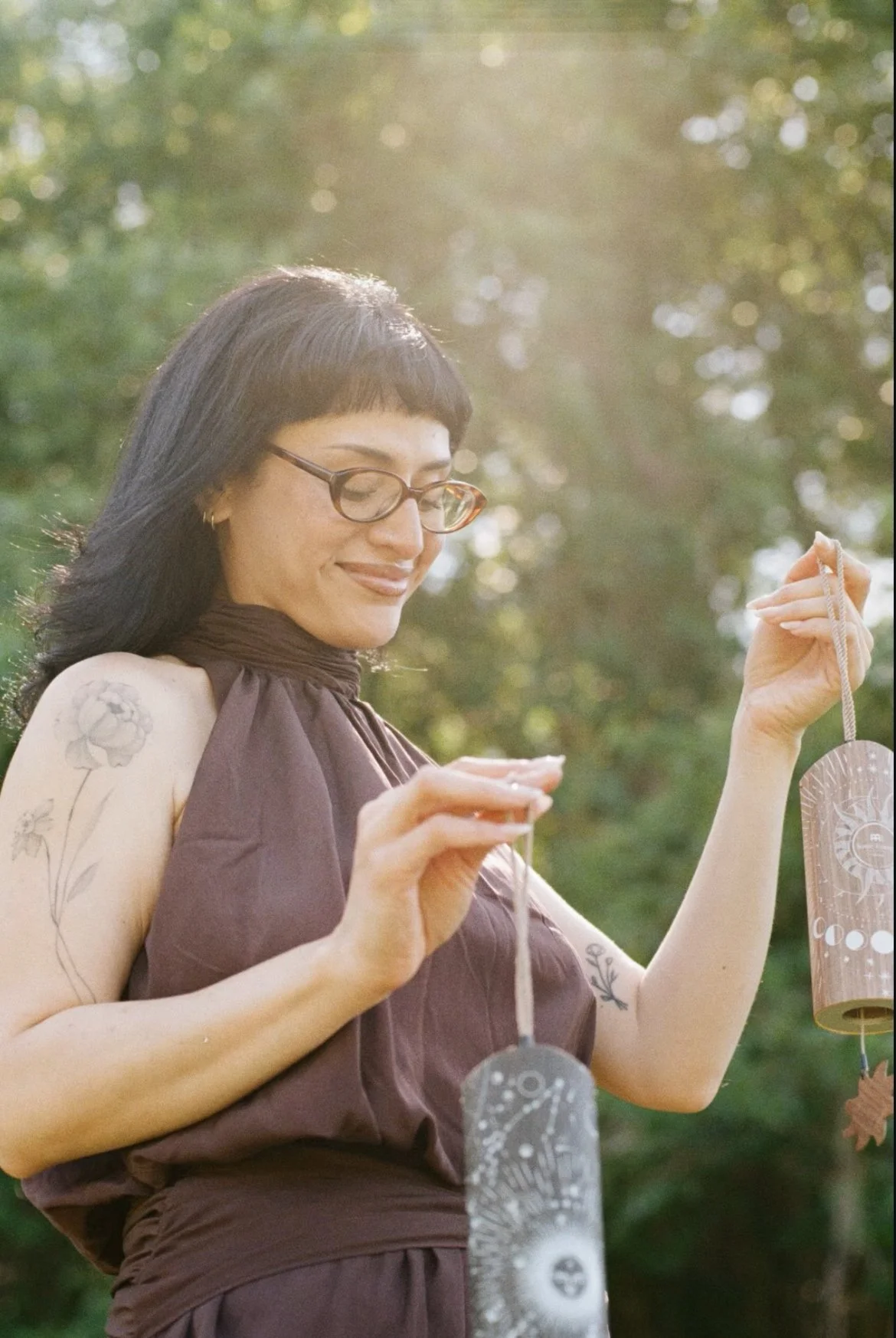 A woman with black hair, glasses, and tattoos on her arms is outdoors, holding and hanging solar-powered lanterns with celestial designs.