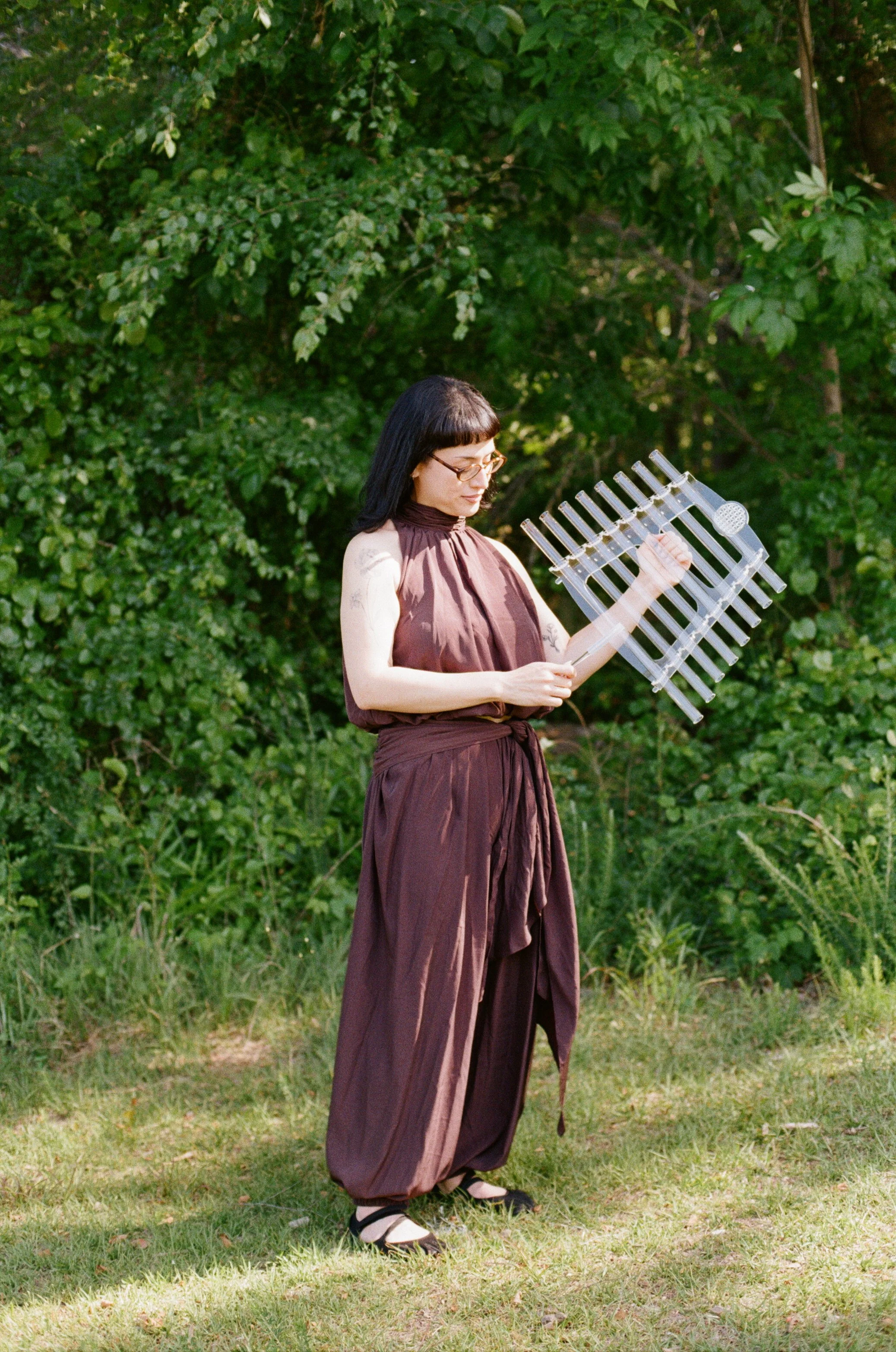 A woman with black hair, glasses, and tattoos on her arms standing outdoors on grass in front of green trees, looking down at a clear xylophone she is holding.