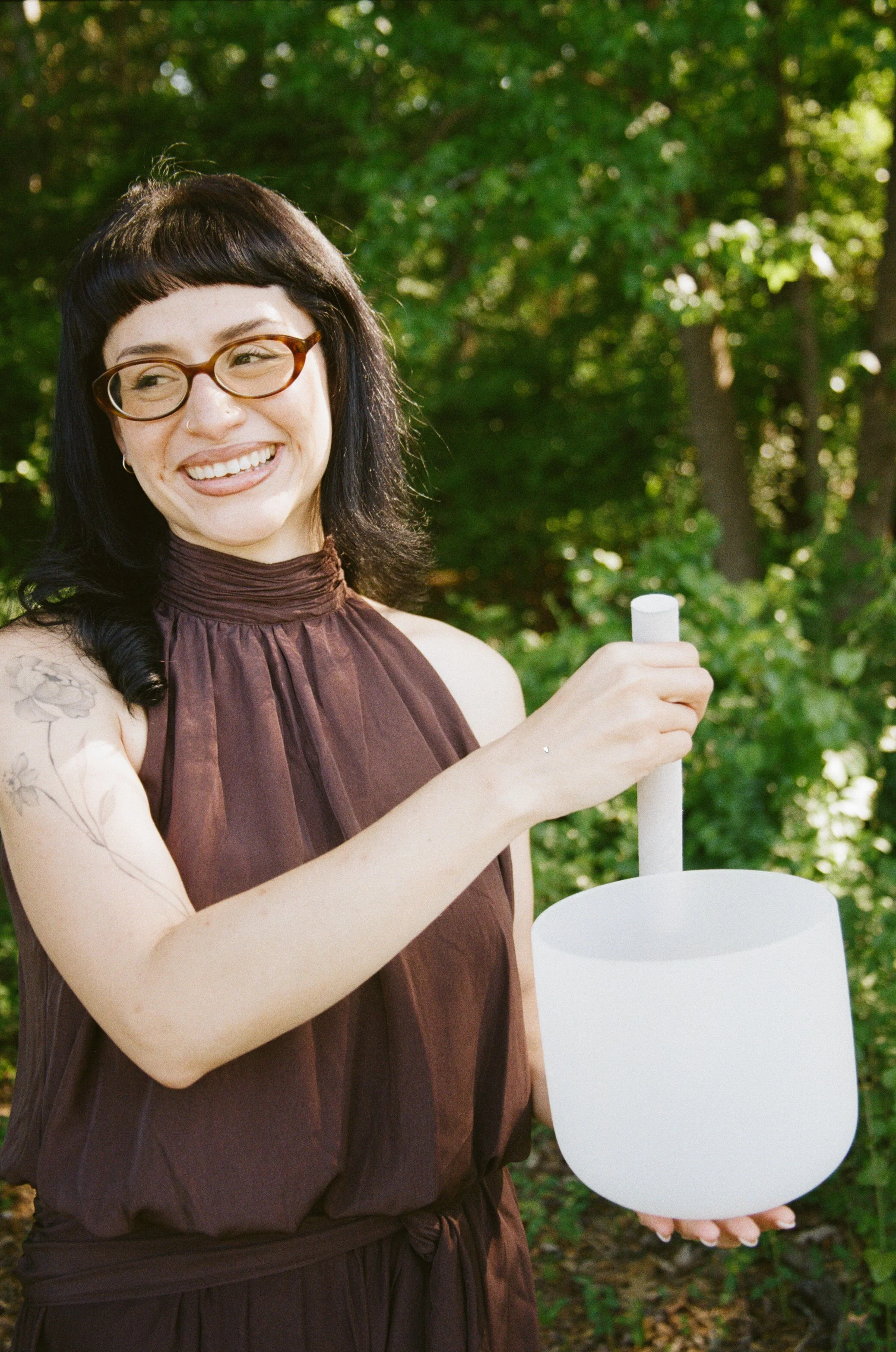 A woman with dark hair, glasses, and tattoos on her arm, smiling outdoors while holding a white drumstick and a white lantern, with green trees in the background.
