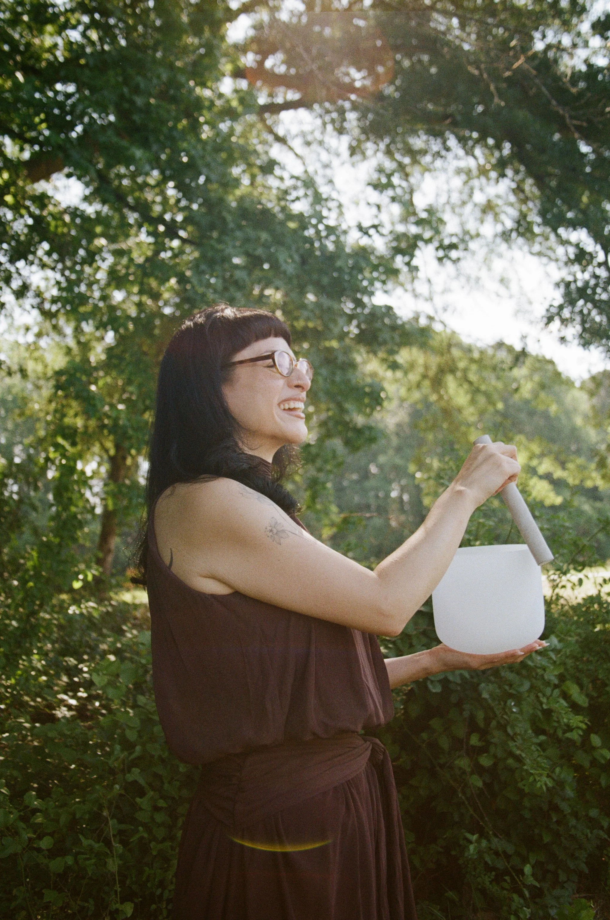 A woman with glasses and dark hair holding a white cylindrical object outdoors surrounded by green trees and foliage.