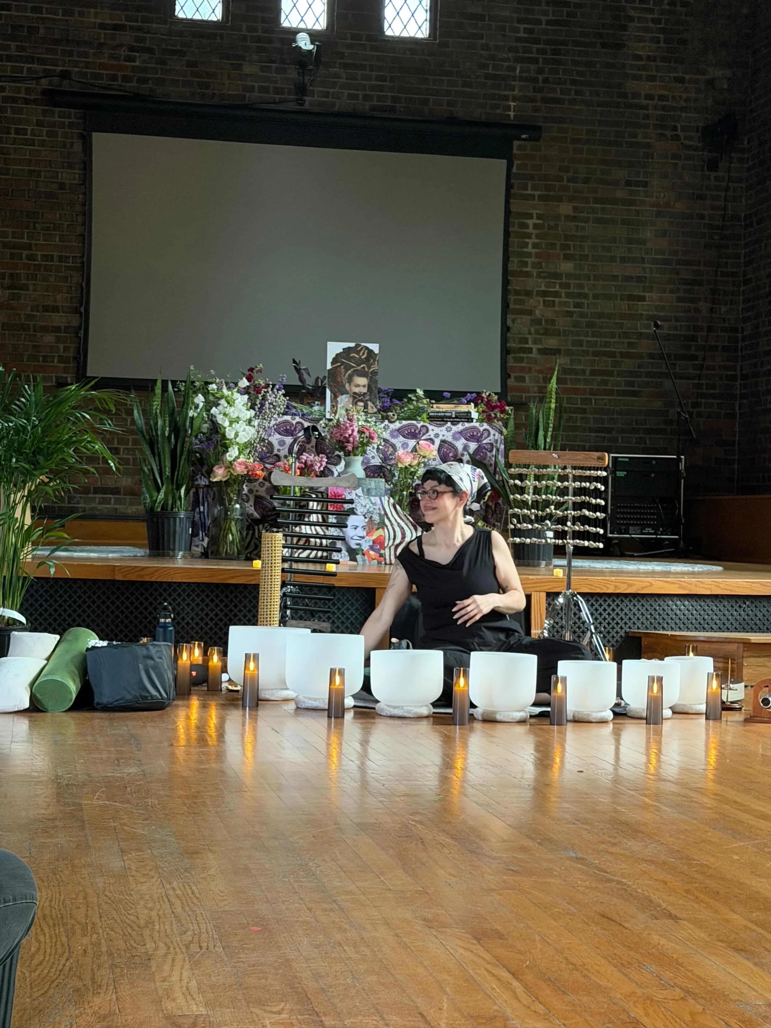 An indoor space decorated for a celebration, with candles, flowers, musical instruments, and a person sitting on the floor. There is a large projection screen on the brick wall, and the setting has a warm, cozy atmosphere.