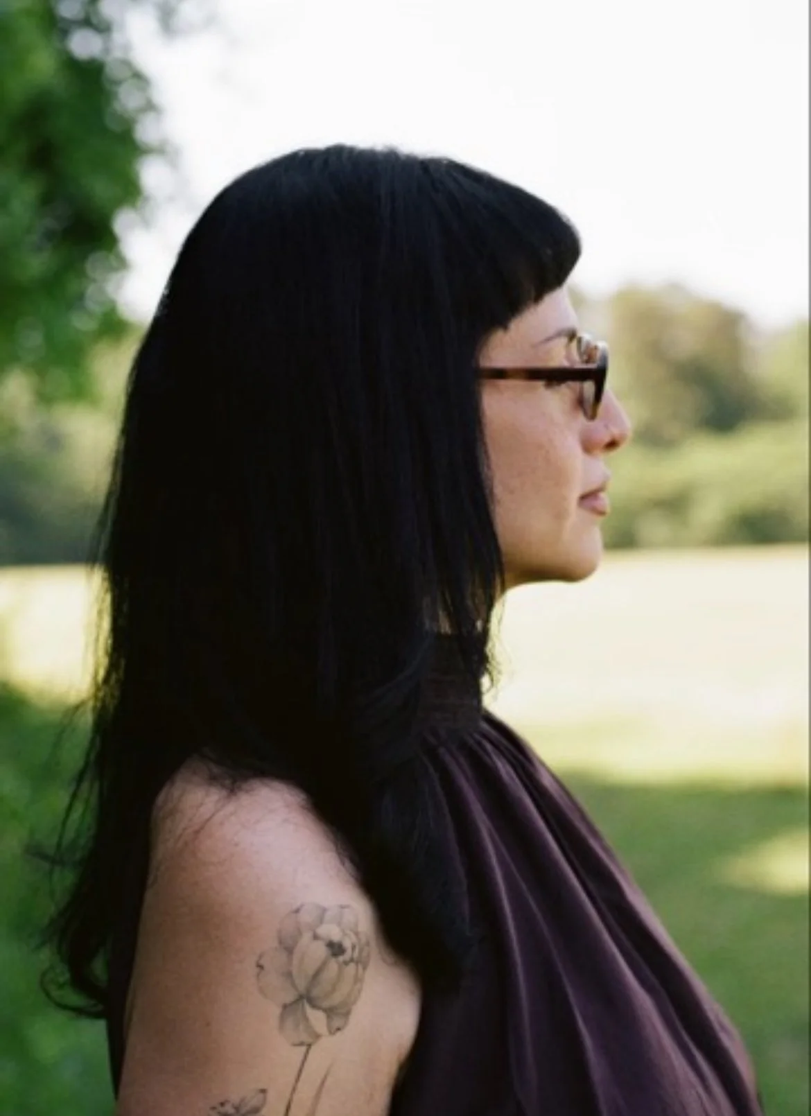 Side profile of a woman with long black hair, glasses, and tattoos on her arm, standing outdoors in a green park.