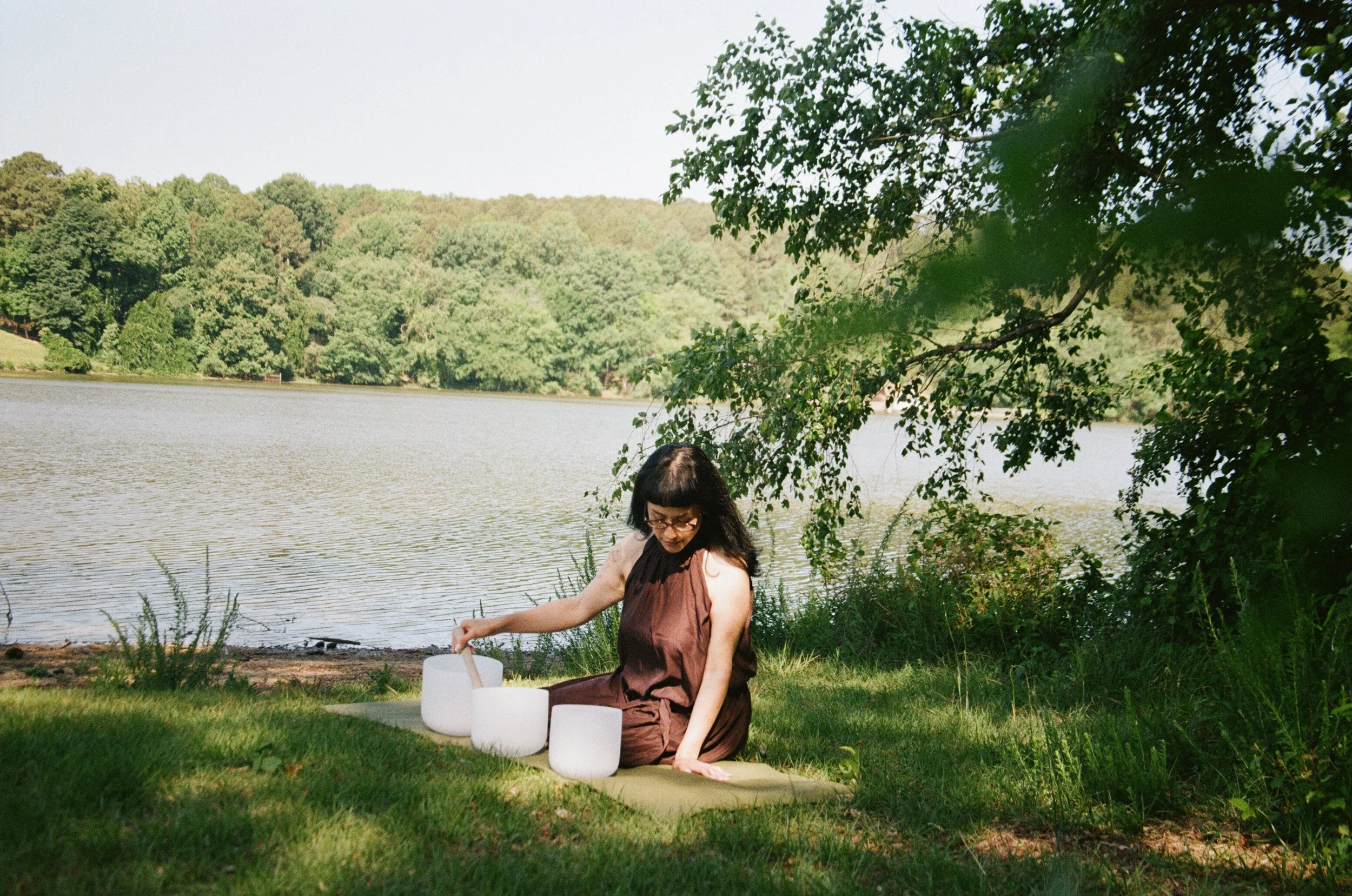 A woman sitting by a lake on the grass during the daytime, surrounded by trees, with sound bowls around her.