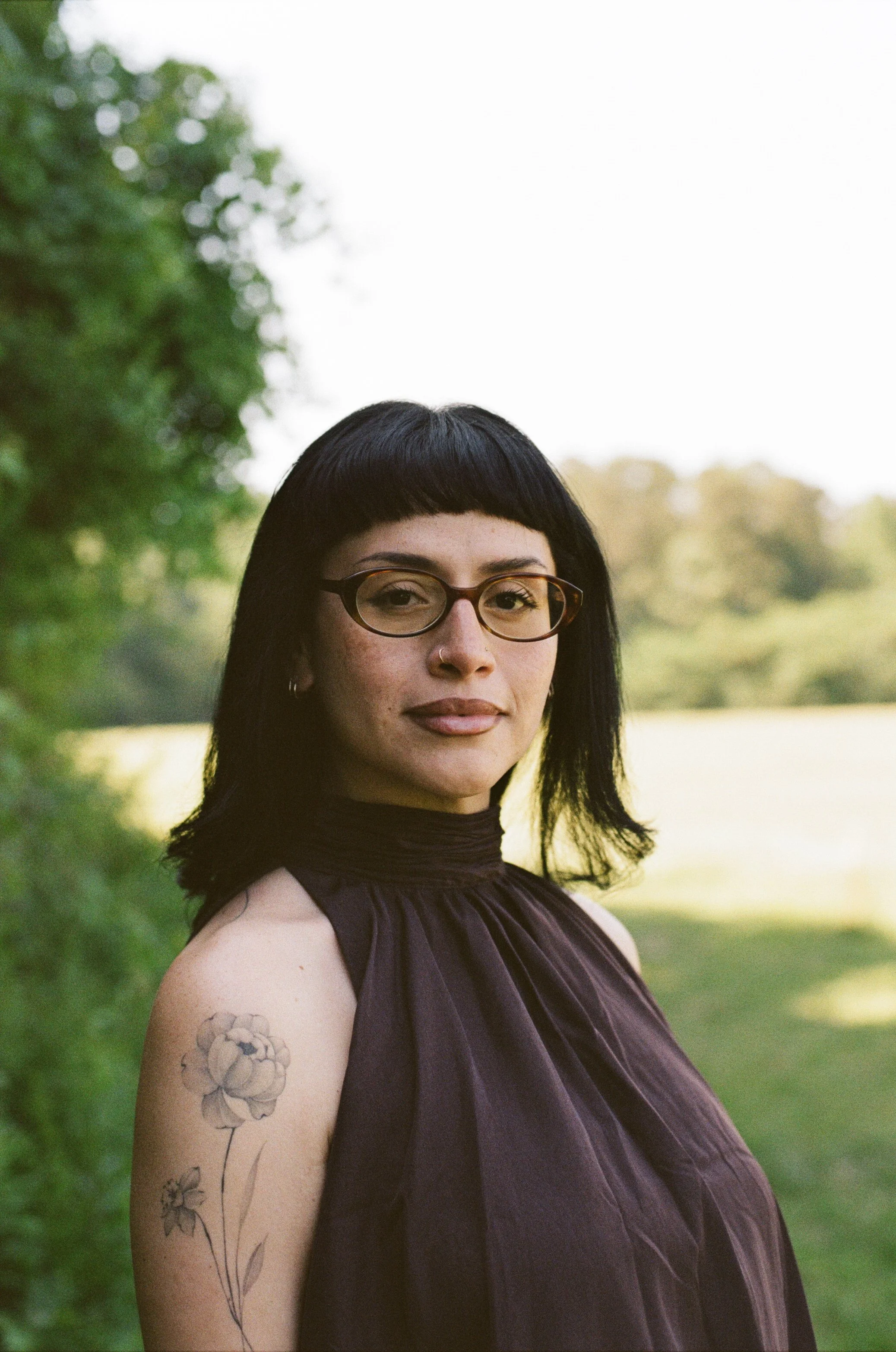 A woman with black hair and glasses standing outdoors in a natural setting, looking into the camera, with trees and a bright sky in the background.