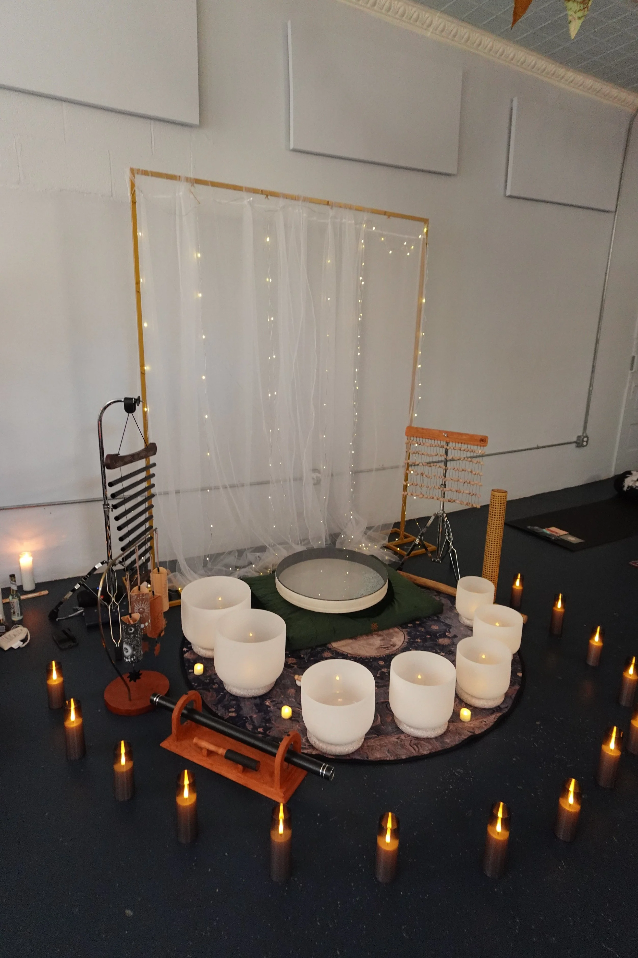 An indoor setup with lit candles and sound instruments arranged in a circle on a dark floor, including singing bowls, with a sheer curtain, fairy lights, and decorative elements in the background.
