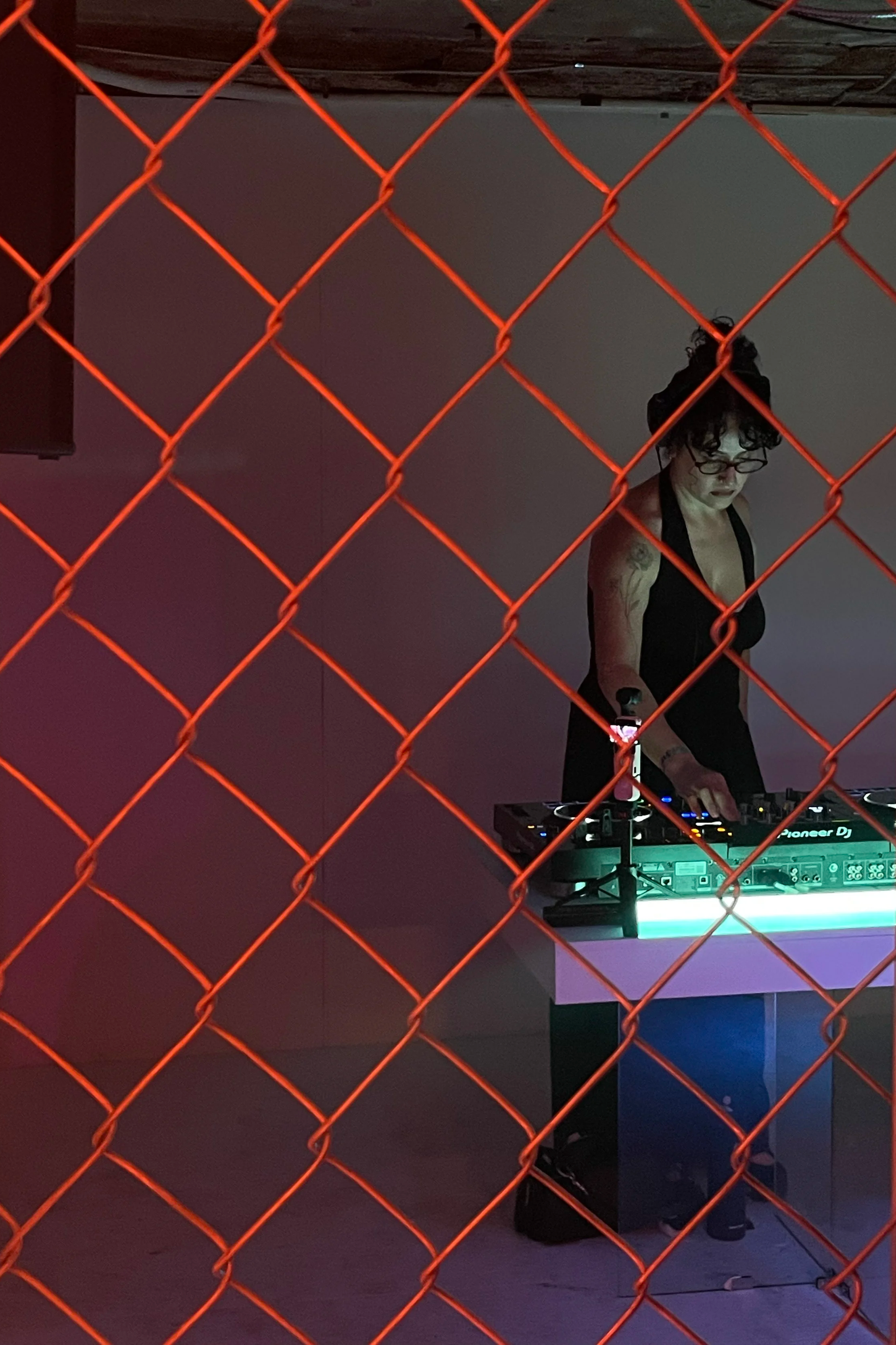 A woman with dark curly hair, glasses, and tattoos on her arms is DJing behind a turntable setup, seen through an orange chain-link fence.