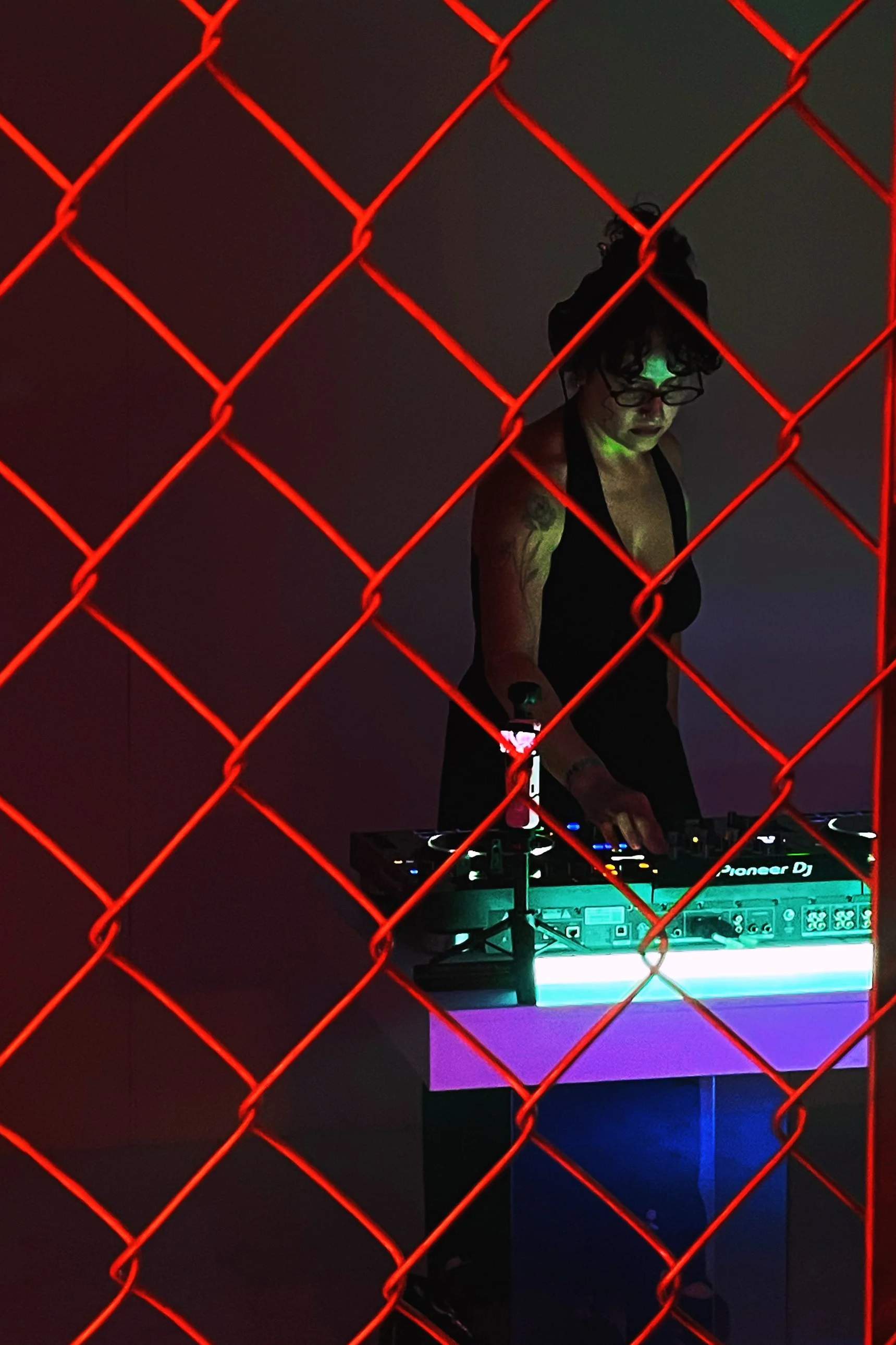 A person with curly hair and glasses, wearing a black top, is DJing behind a glowing DJ controller. The scene is viewed through a red chain-link fence with dark surroundings.