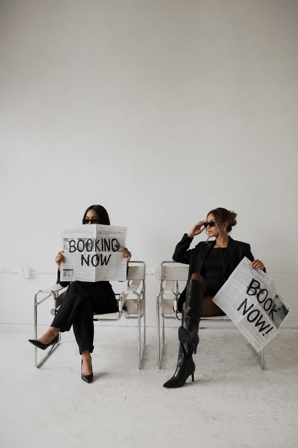 Two women sitting on chairs against a plain white wall, wearing black outfits and sunglasses. Both are holding newspapers with large printed text reading 'BOOKING NOW', and one woman is reading the newspaper while the other is adjusting her sunglasses.