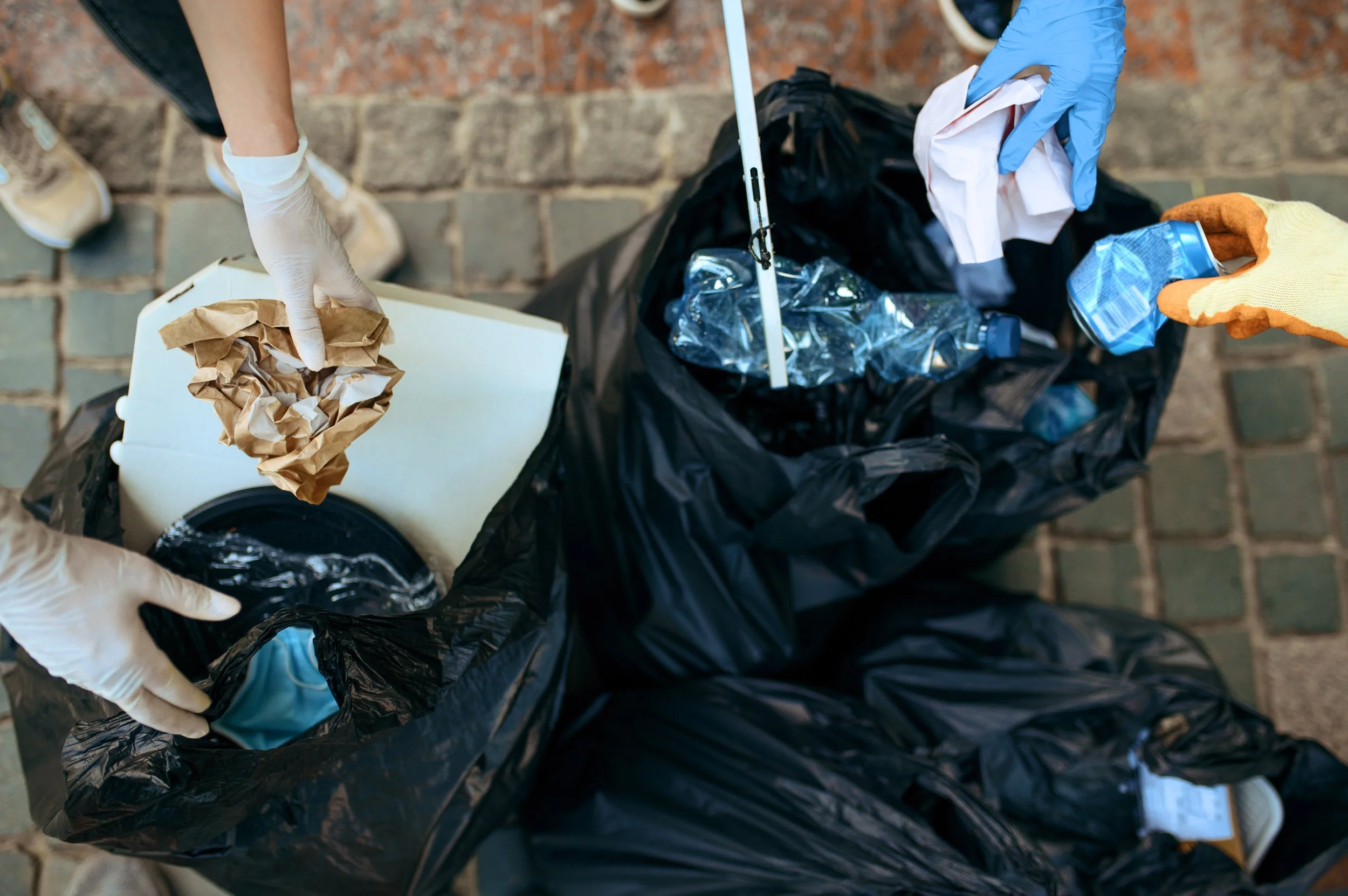 Two people wearing gloves are trash picking, collecting litter such as crumpled paper and plastic bottles into a black trash bag.