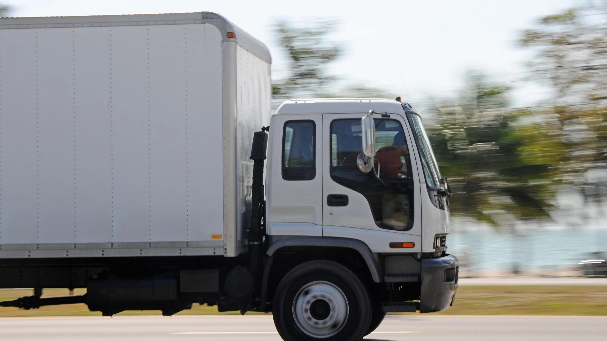 A white delivery truck driving at high speed on a road with blurred trees in the background.