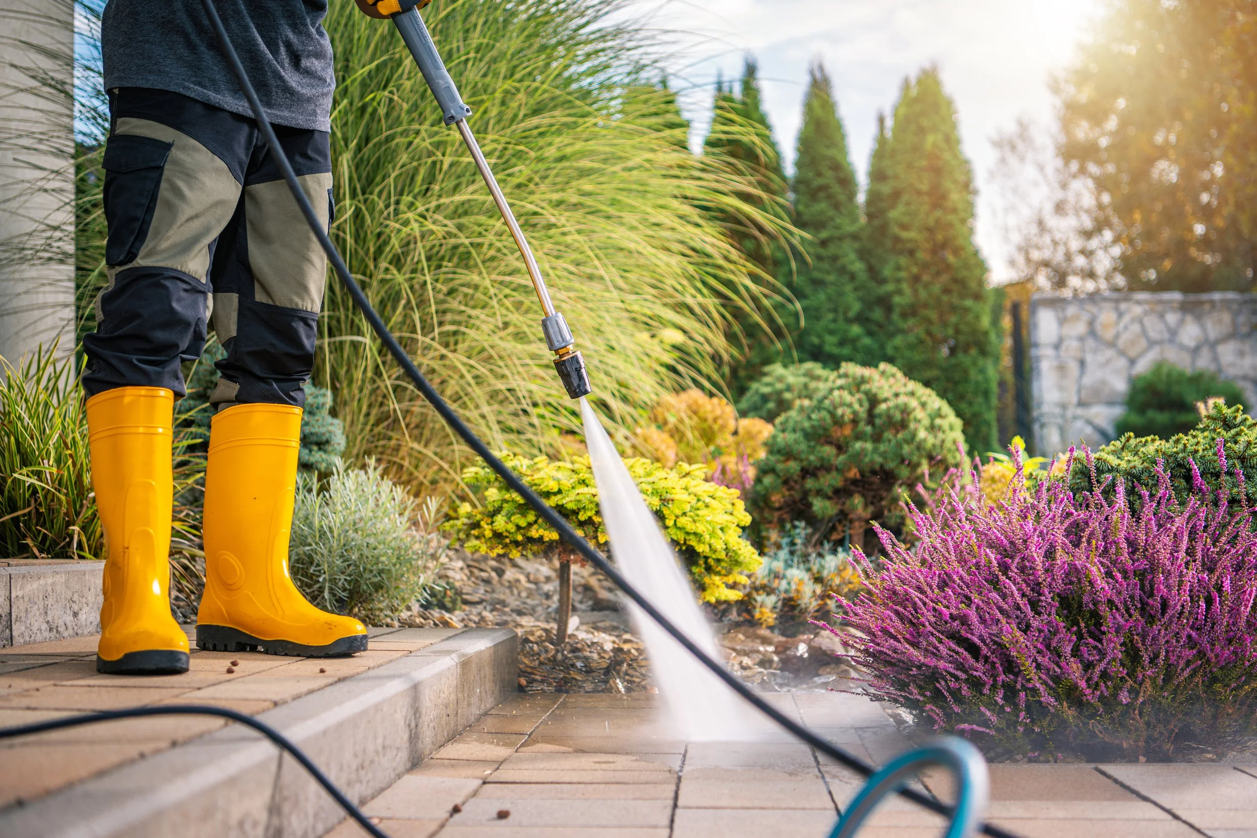 Person in yellow rain boots watering plants in a garden with colorful flowers and tall green trees in the background.