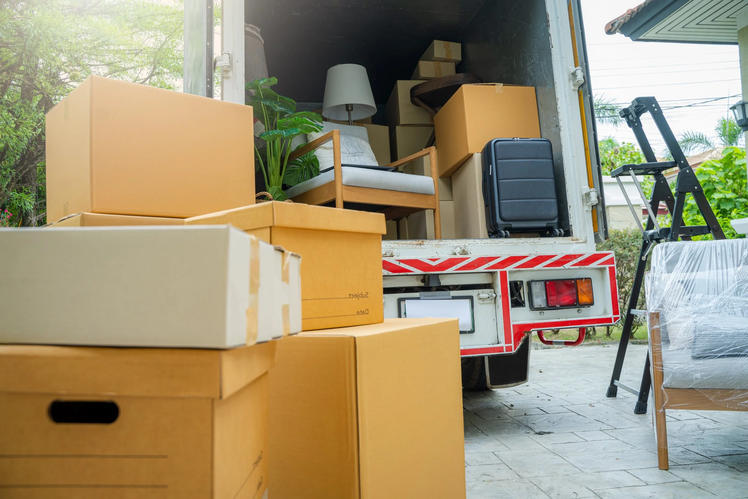 Moving truck filled with boxes, furniture, and household items, with some boxes outside the truck and folding chairs nearby, on a paved area outdoors.
