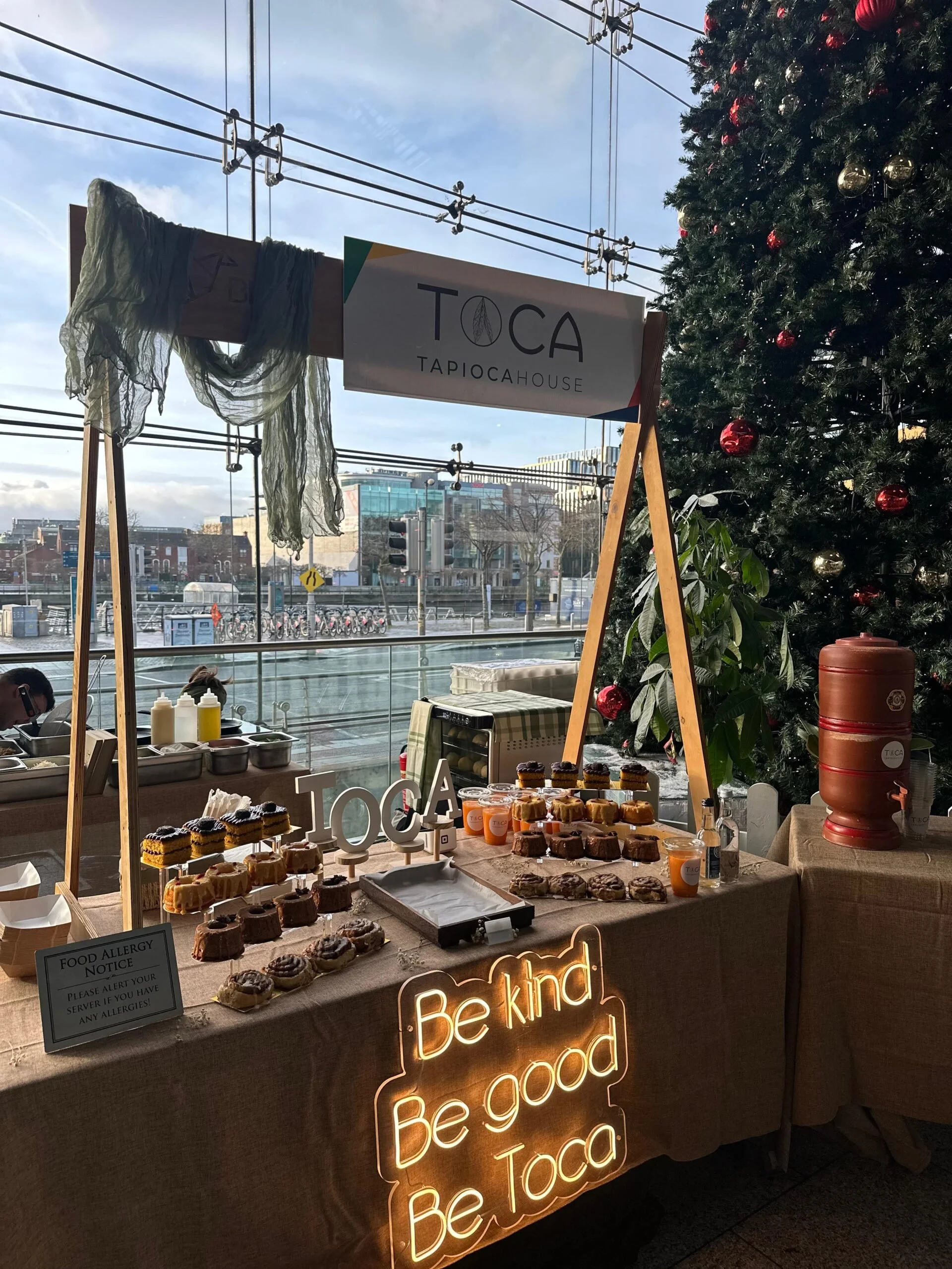 Decorated Christmas tree next to a dessert table with assorted baked goods and a neon sign that says, "Be kind Be good Be Toca."
