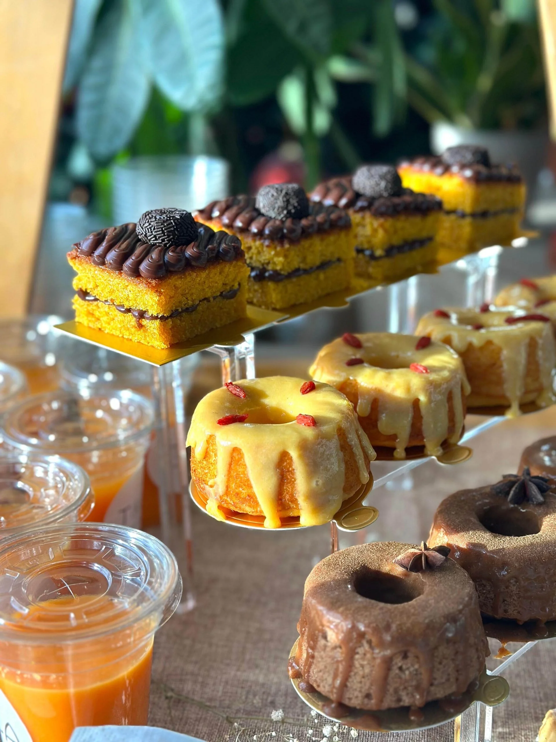 Close-up of assorted desserts on a tiered stand, including yellow cake slices with chocolate and cookie decorations, and small cups of orange-colored drink on the side.