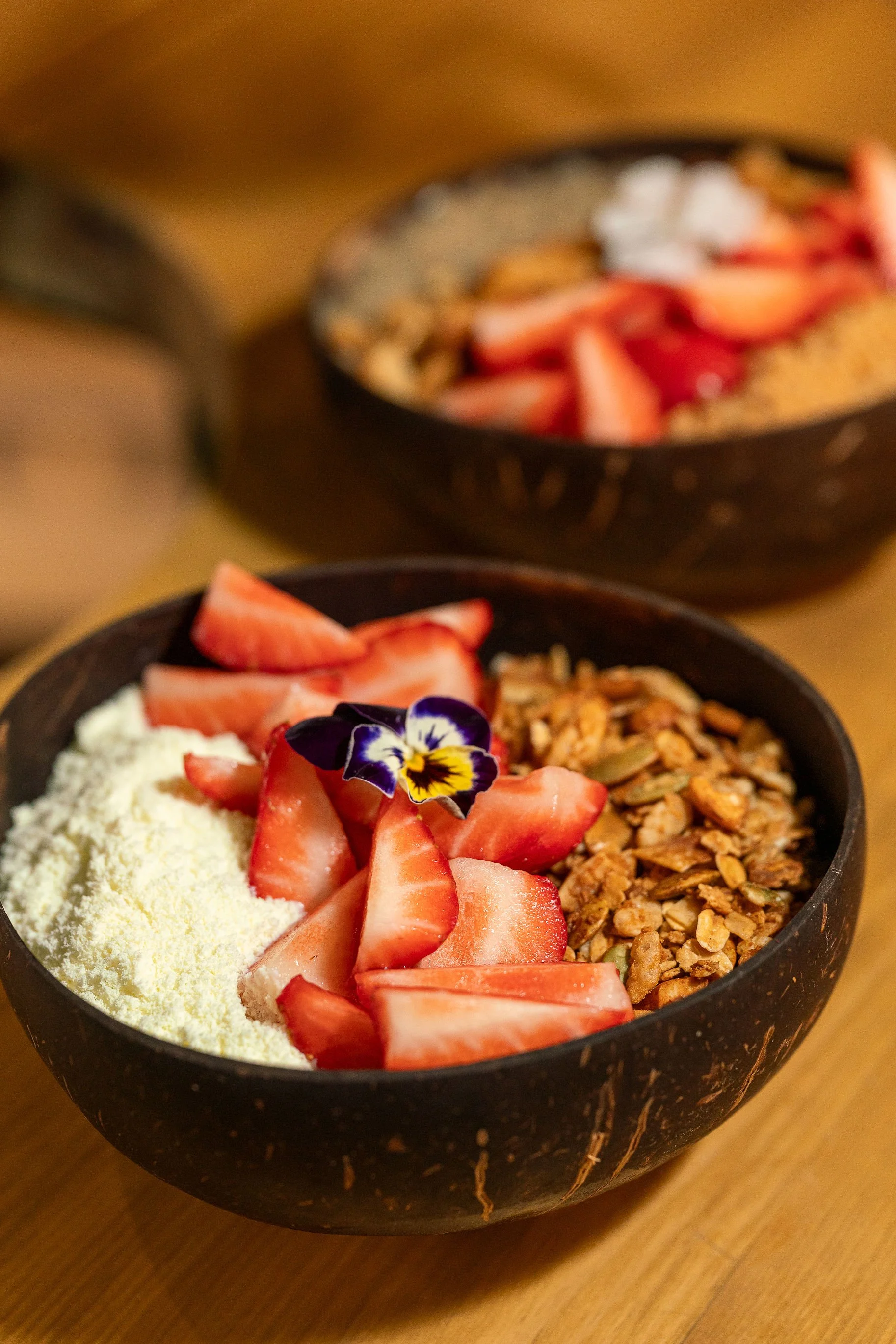 Close-up of a Brazilian Style Açaí bowl with strawberries, powered milk, homemade granola, condensed milk and edible flower petals on top, placed on a wooden surface with another similar bowl in the background.