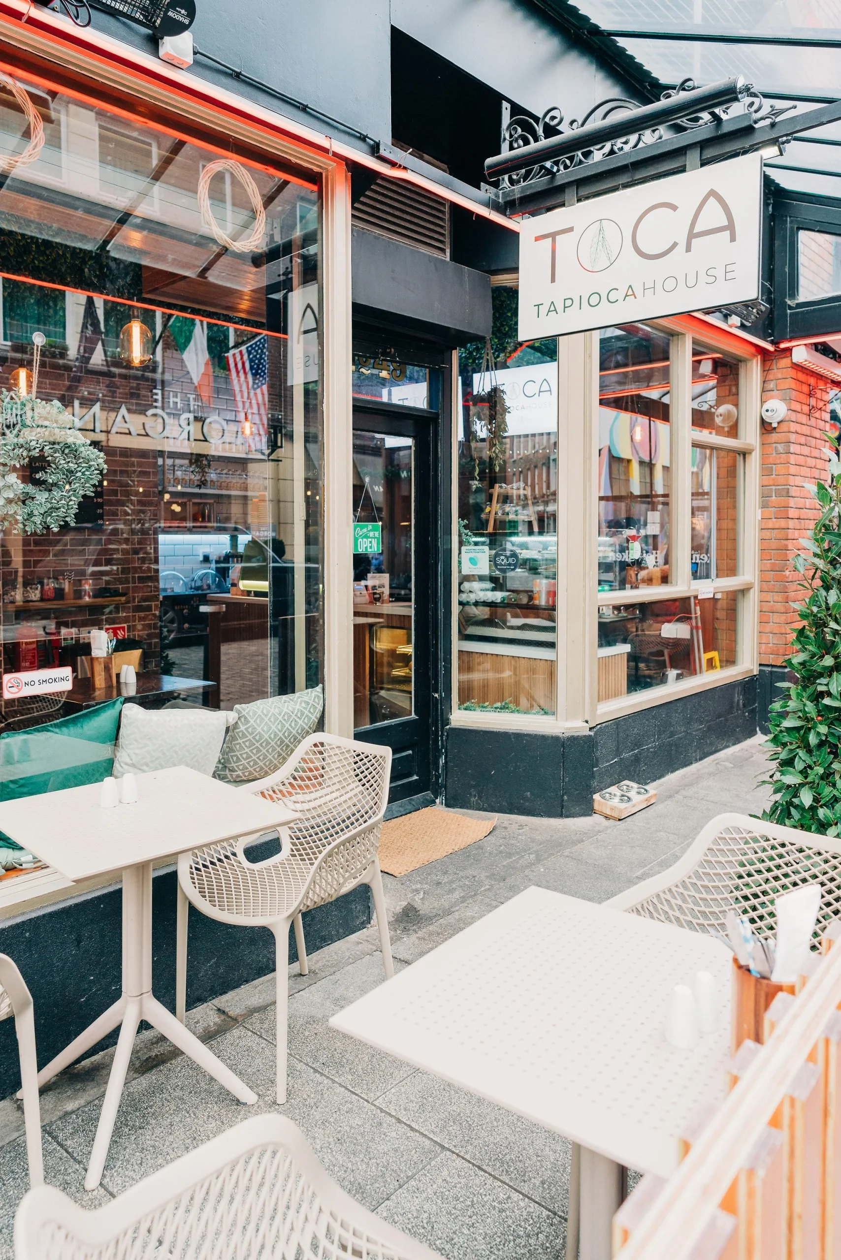 Outdoor patio seating area with white chairs and tables in front of a restaurant named Toca Tapioca House. The restaurant has large glass windows, interior warm lighting, and a sign hanging outside. The sidewalk is paved, and there are some plants along the edge.