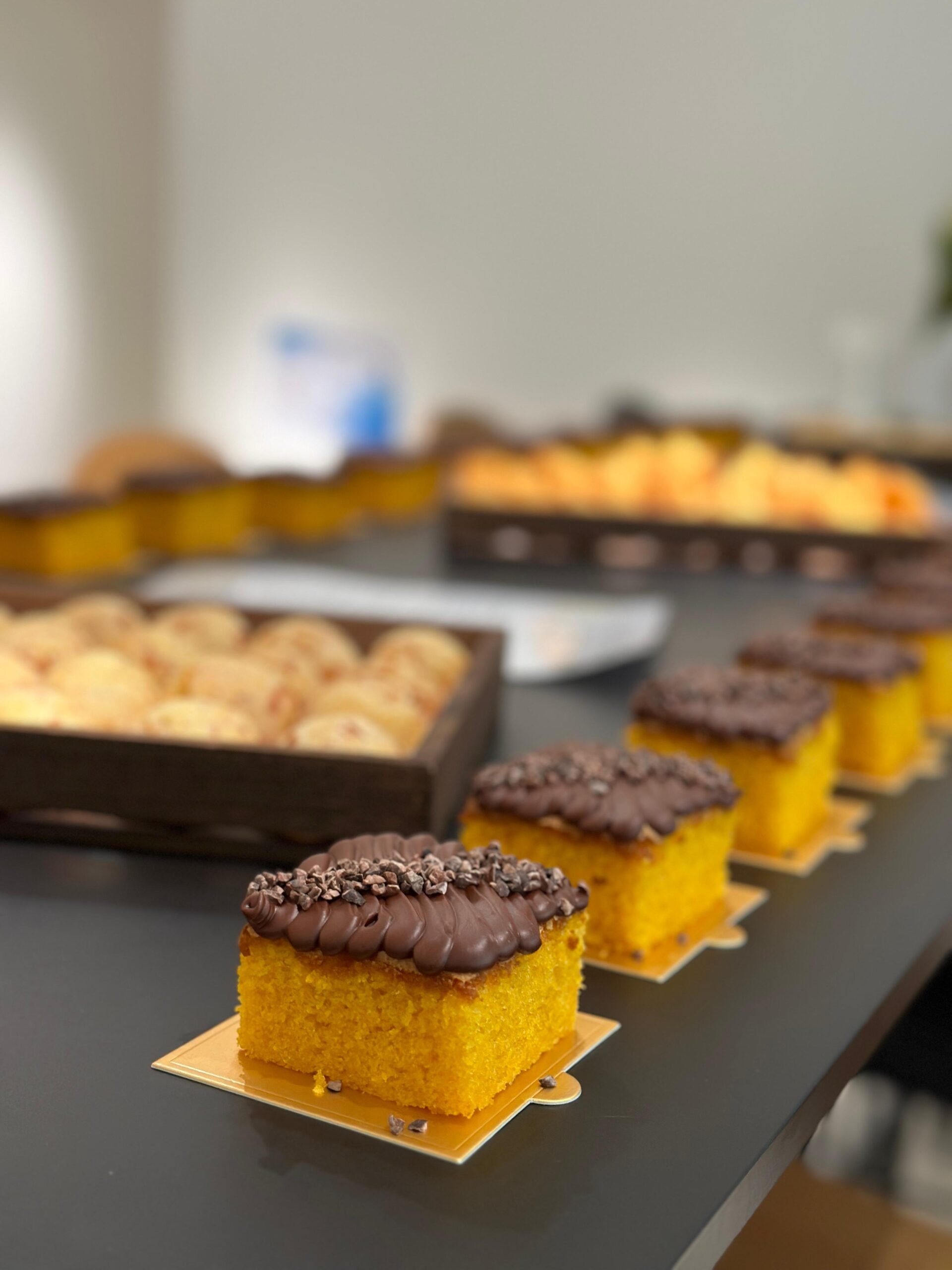 Close-up of a yellow cake square topped with chocolate frosting and chocolate sprinkles, in the foreground. In the background, blurred trays of various desserts, including cookies and other pastries, on a dark table.