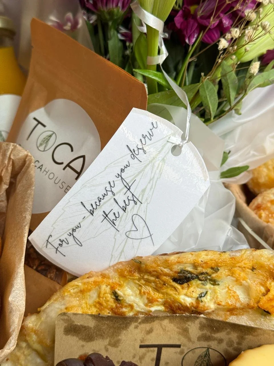 Close-up of a slice of pizza with cheese and herbs, surrounded by flowers, a paper bag, and a note that reads 'Tay you because you deserve the best' with a heart drawing.