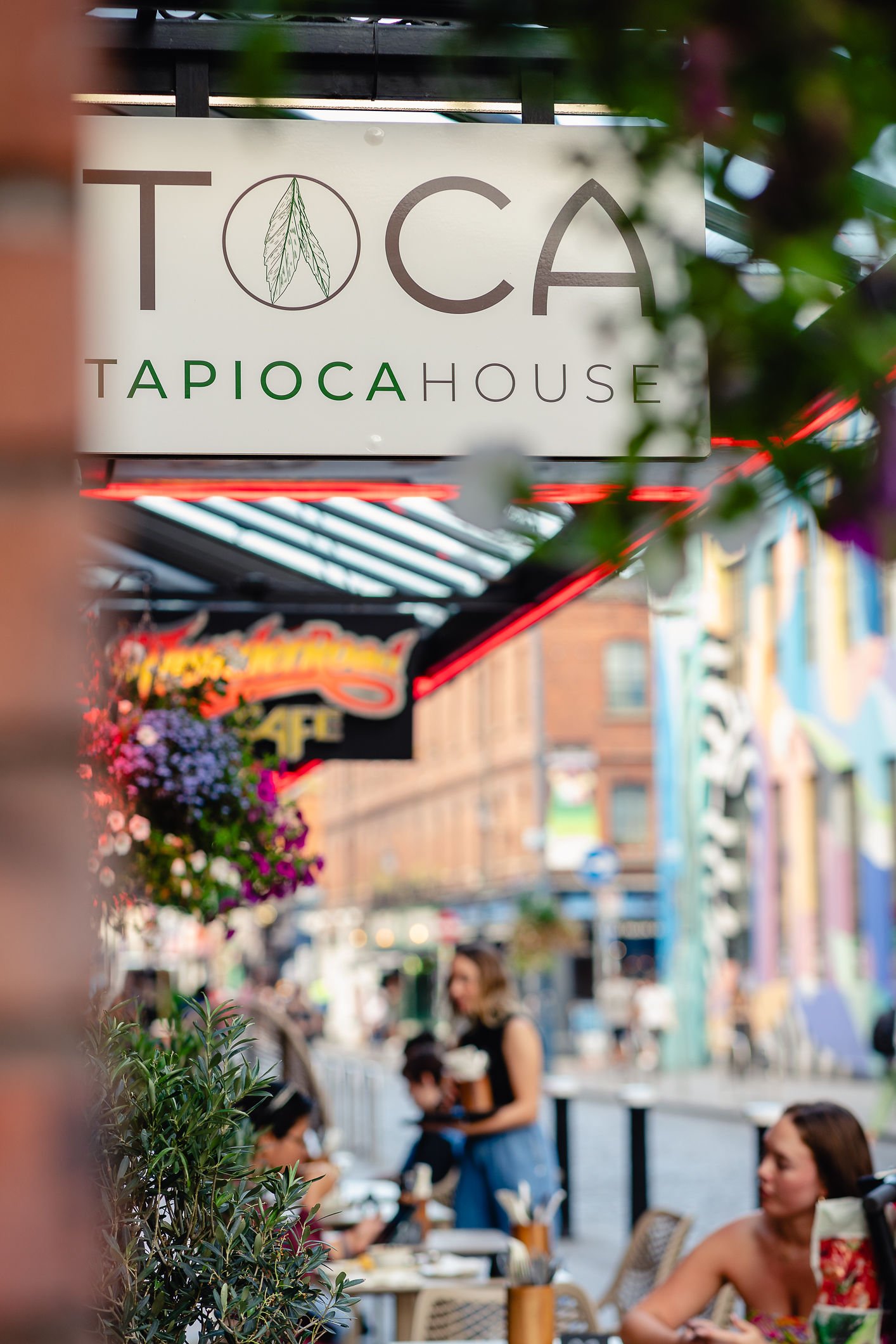 Sign for Toca Tapioca House shop with people dining outdoors in an urban setting, greenery, and colorful floral arrangements