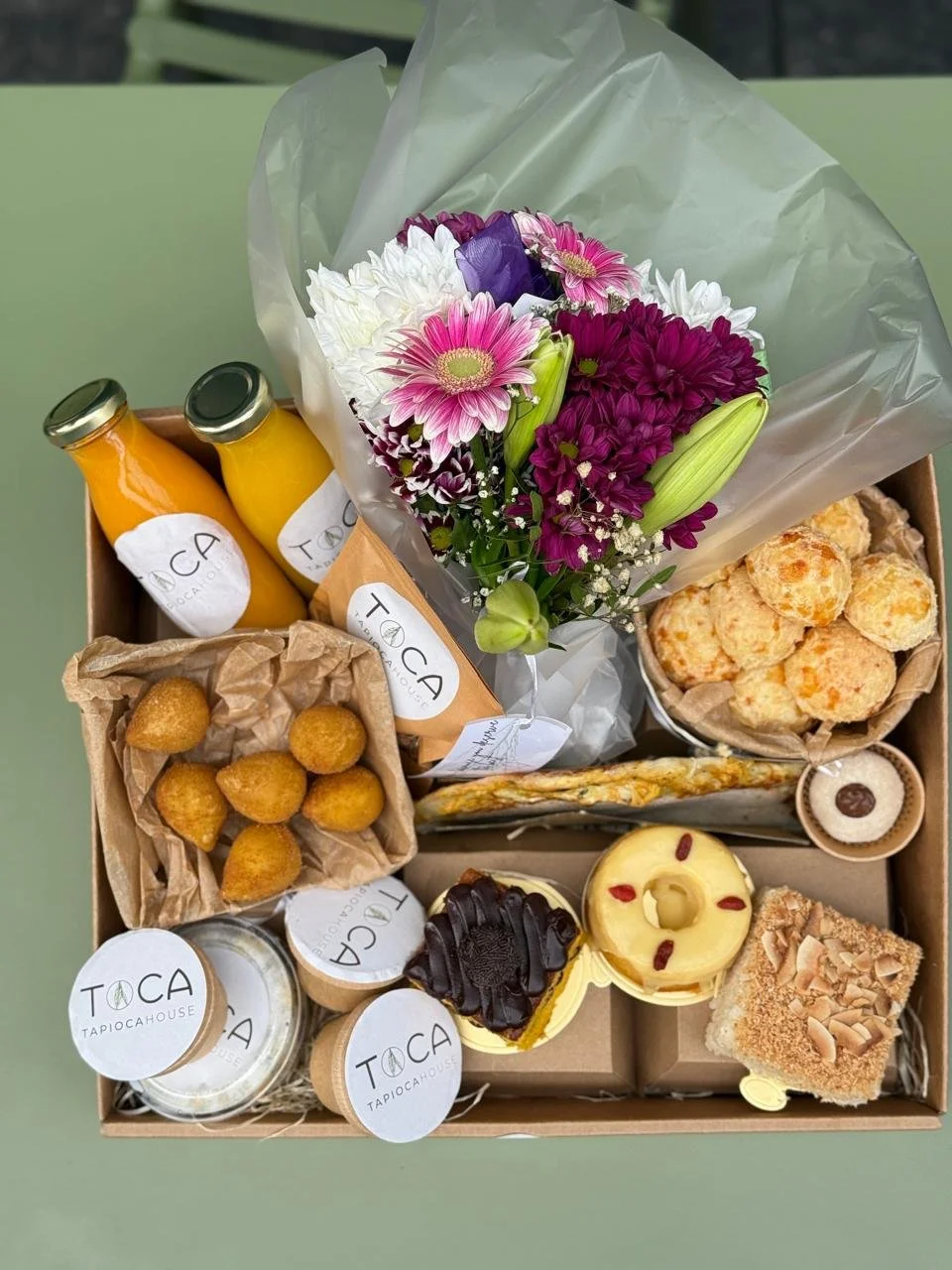An assorted gift box containing a bouquet of colorful flowers, two bottles of orange and yellow juice, fried foods, small desserts, and other snacks from Toca Tapioca House.