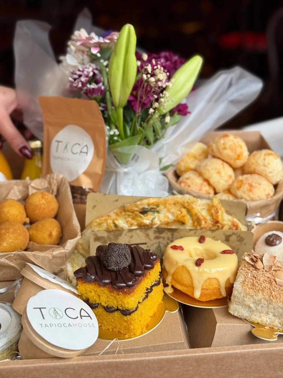 An assortment of desserts including a yellow cake with chocolate and Oreo toppings, a glazed donut with red sprinkles, coconut-coated pastry, and a slice of vegetable quiche, along with a bouquet of lilies and other flowers, and a small package labeled 'Toca' on a table.