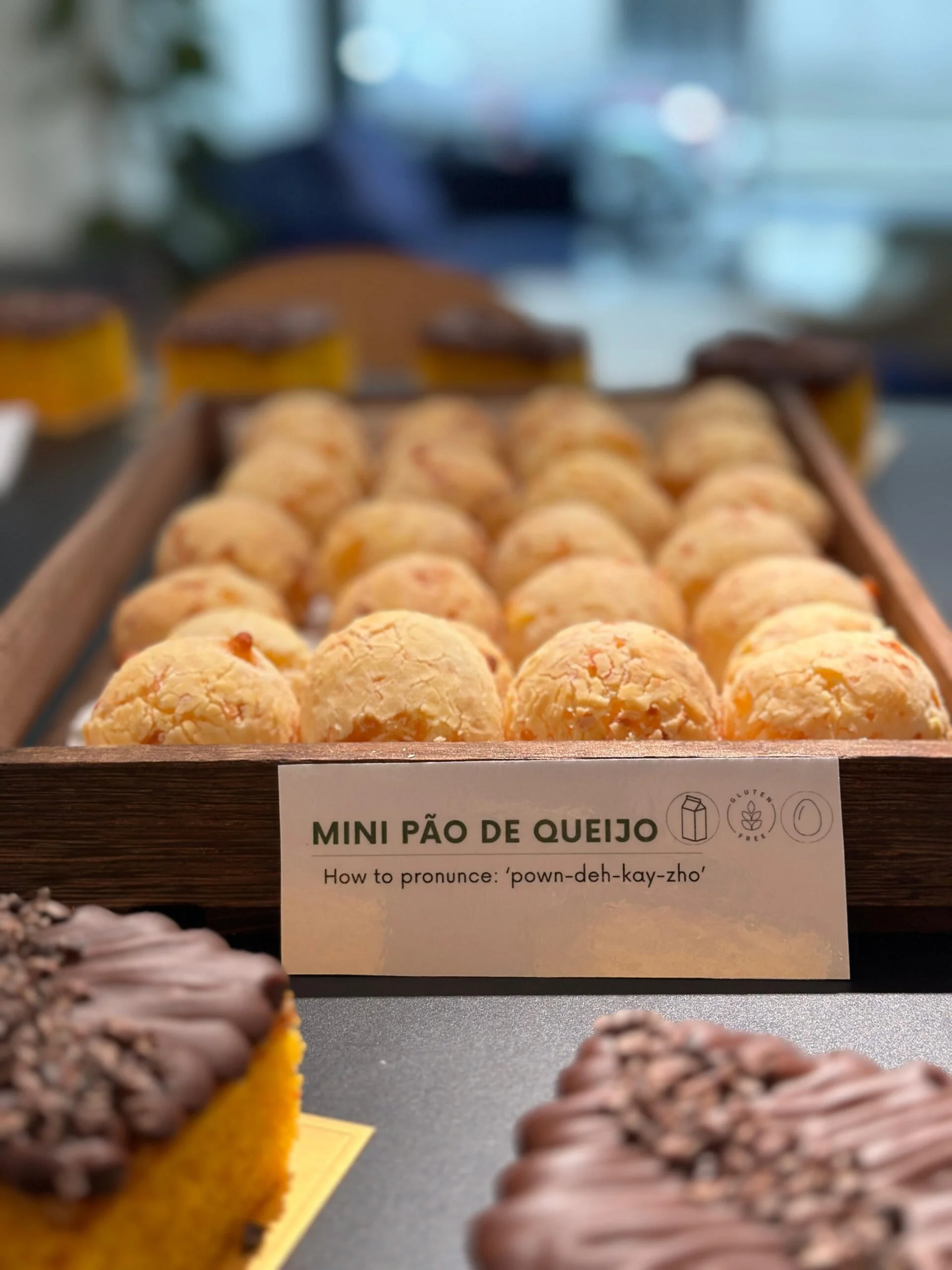 Display of mini cheese bread and other baked goods at a bakery with a sign explaining how to pronounce 'Mini Pão de Queijo'.