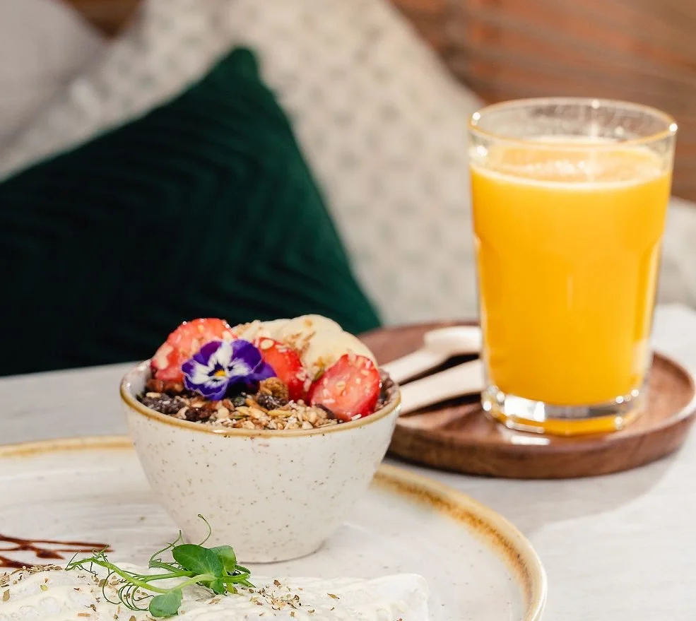 A bowl of açaí topped with granola, strawberries, and an edible flower. A glass of orange juice on a wooden tray next to a fork and spoon. A green cushion in the background.
