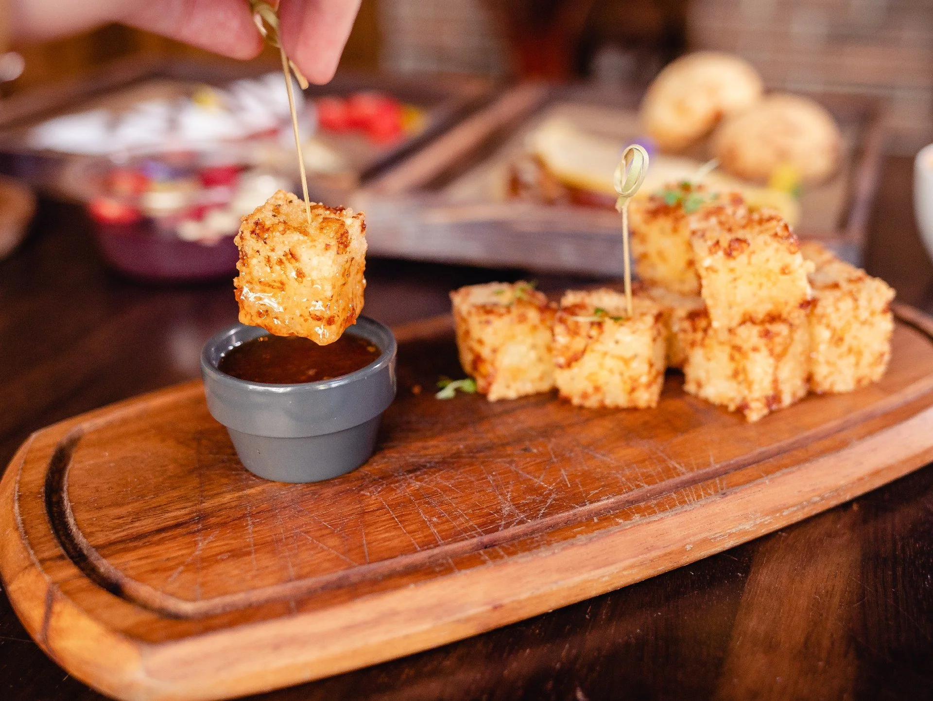 A hand is dipping a piece of crispy tapioca dice into a small bowl of sweet and sour sauce on a wooden serving platter with crispy pieces of fried tapioca dice