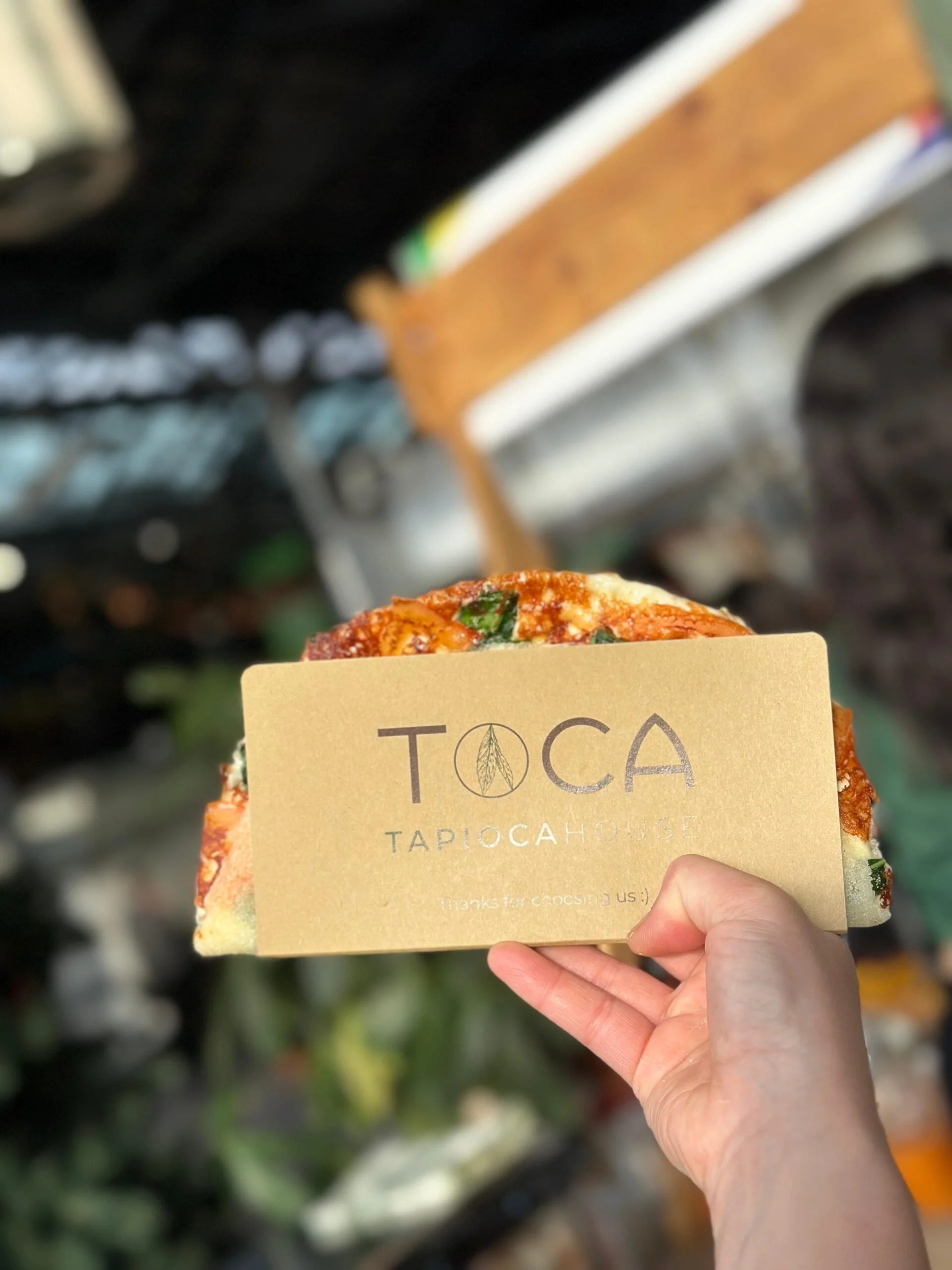 A hand holding a tan business card with the logo and text 'TOCA TAPIOCA HOUSE' in front of blurry shelves of baked goods.