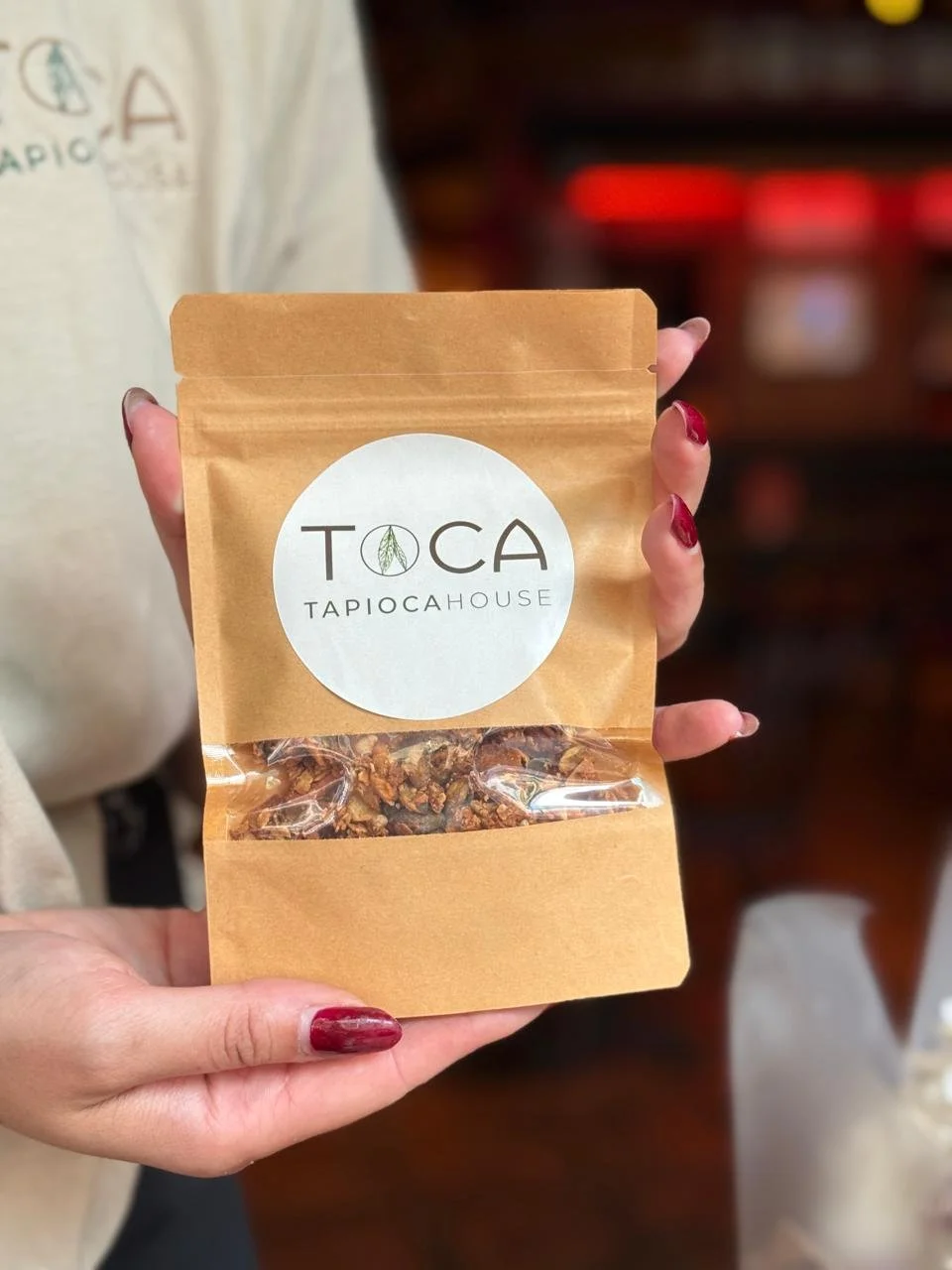 Person holding a brown paper bag with a white circular logo reading TOCA TAPIOCAHOUSE, showing granola or trail mix inside.
