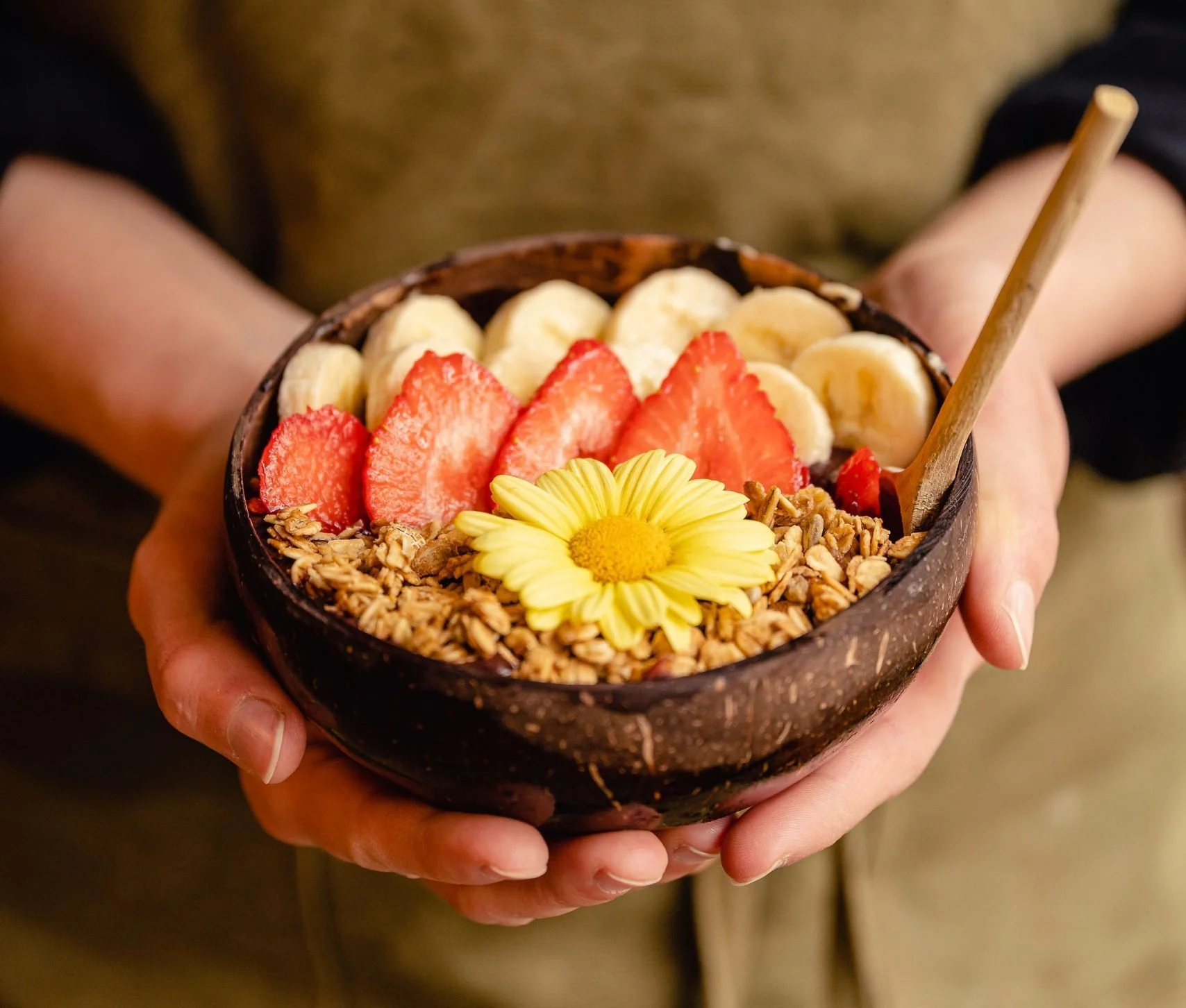 Person holding a coconut shell bowl filled with Açaí, sliced bananas, strawberries, granola, and a yellow flower, with a wooden spoon.
