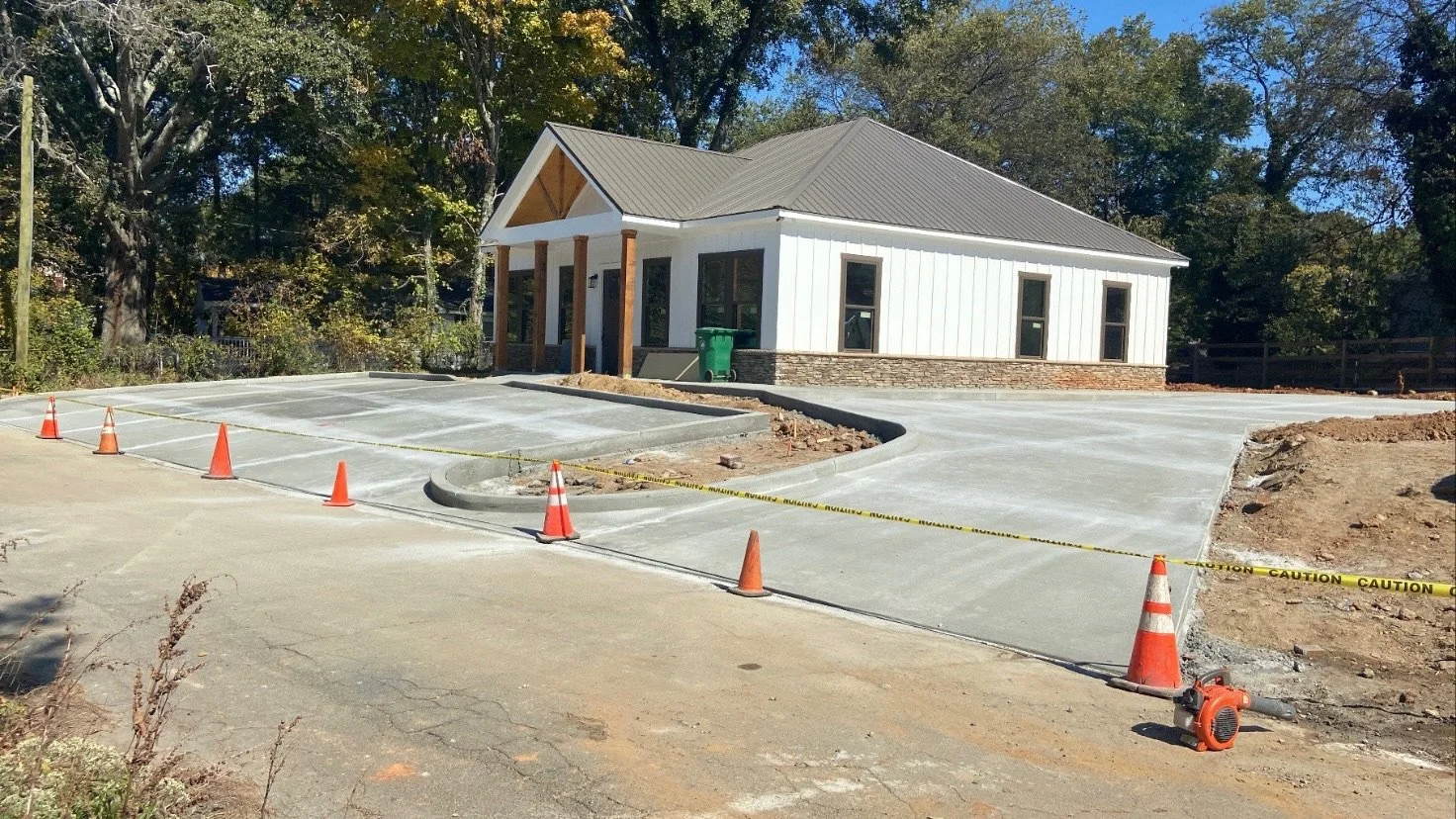 Construction site with a house and freshly poured concrete driveway, marked by orange cones and caution tape, with trees in the background.