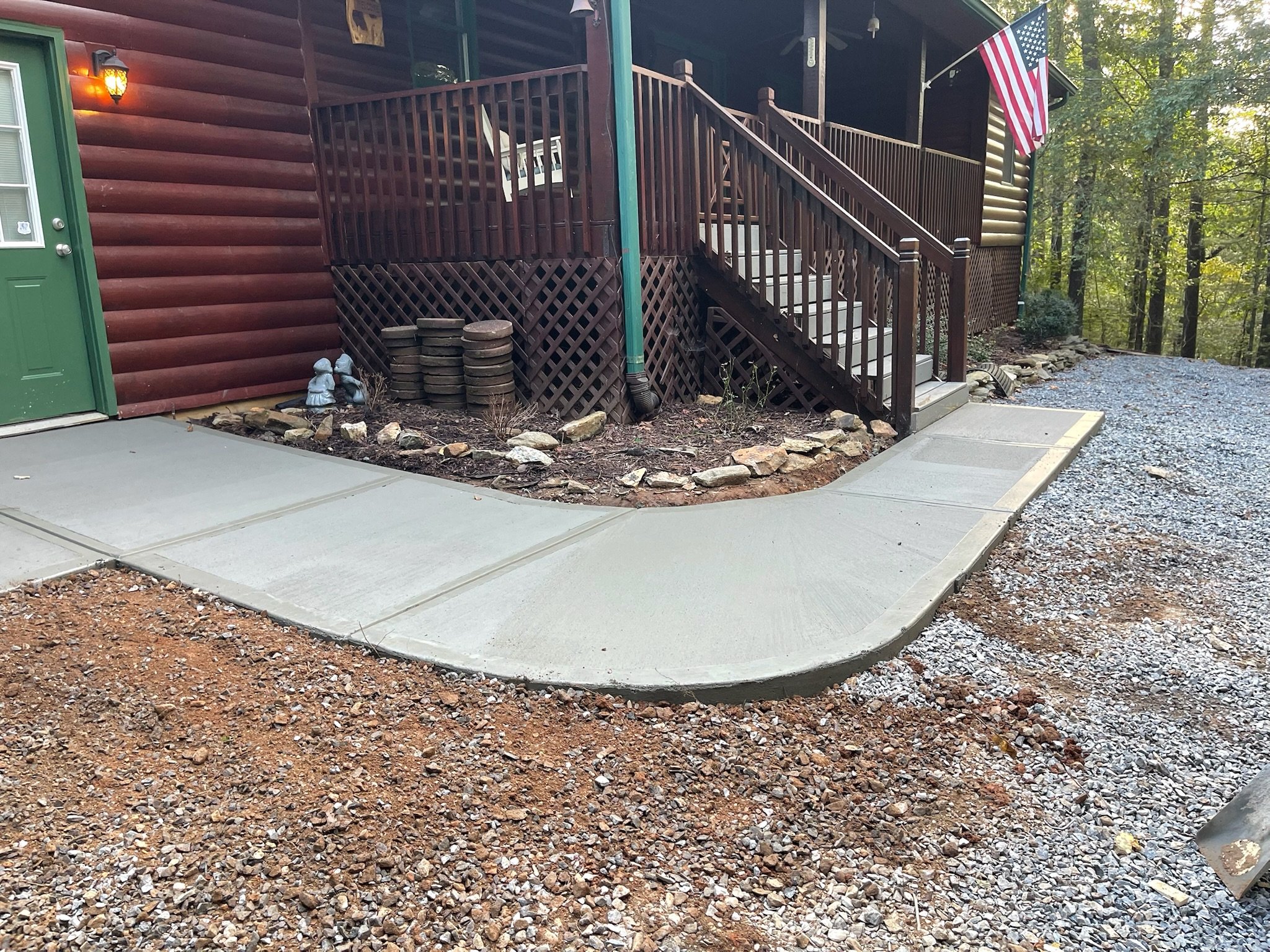 Newly poured concrete sidewalk in front of a house with a brown wooden porch and stairs, a green door, a flag, and decorative garden statues.