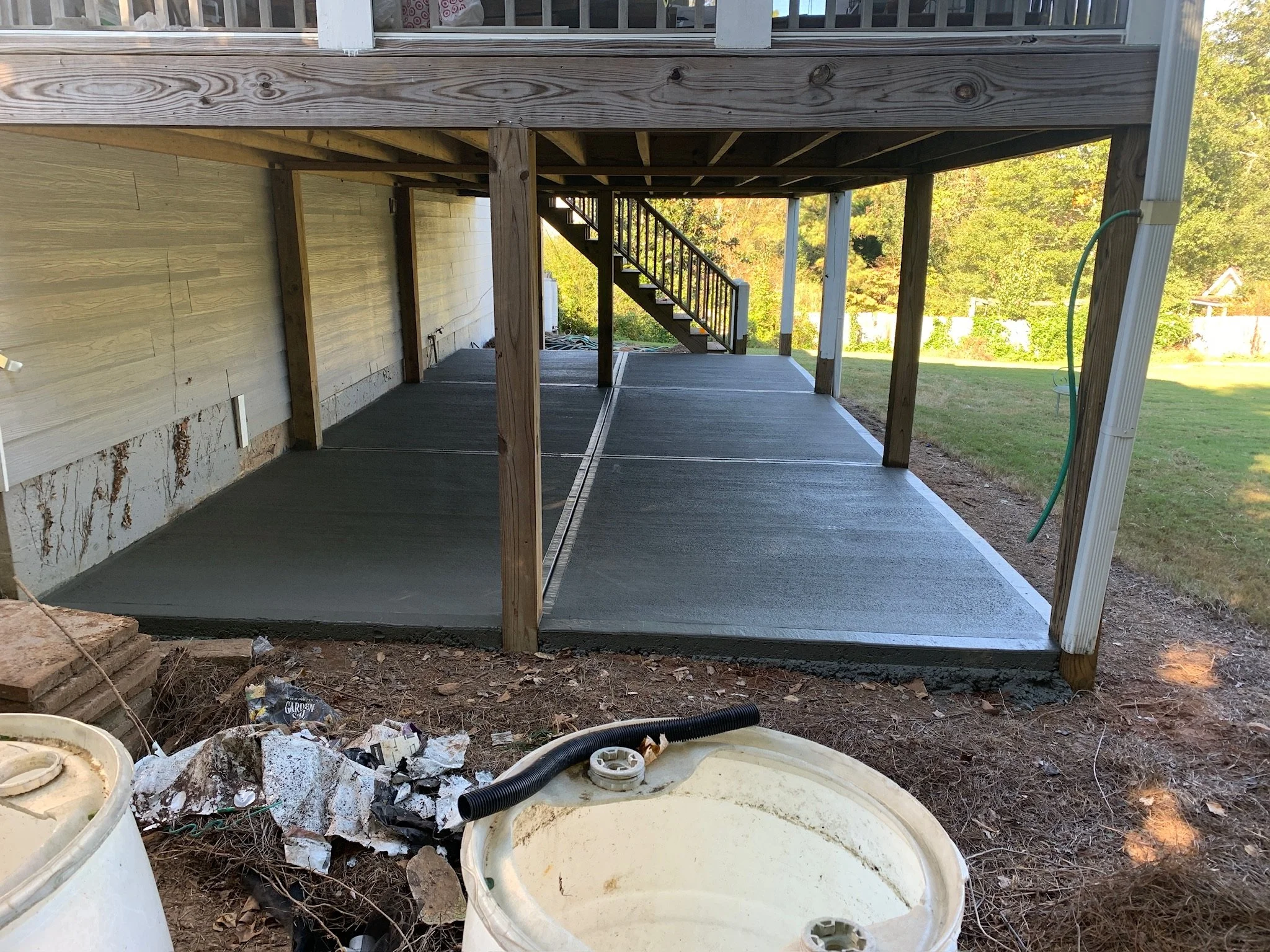 View under a house with a new wooden deck and stairs, a yellow wall, and construction debris on the ground, including white plastic containers, pipes, and rocks.