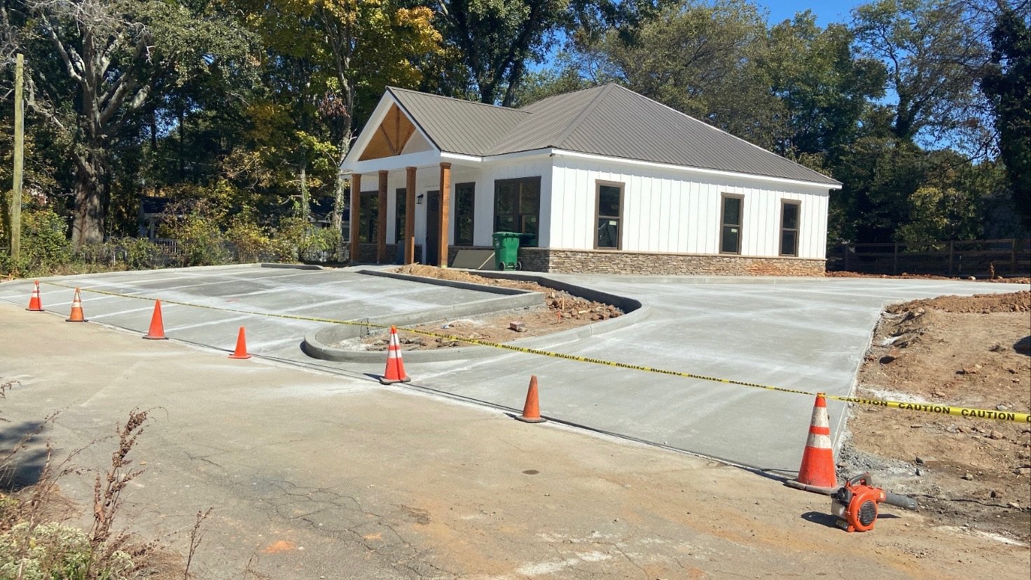 Construction site with a newly paved parking lot and a small white building with a gray roof, surrounded by trees.