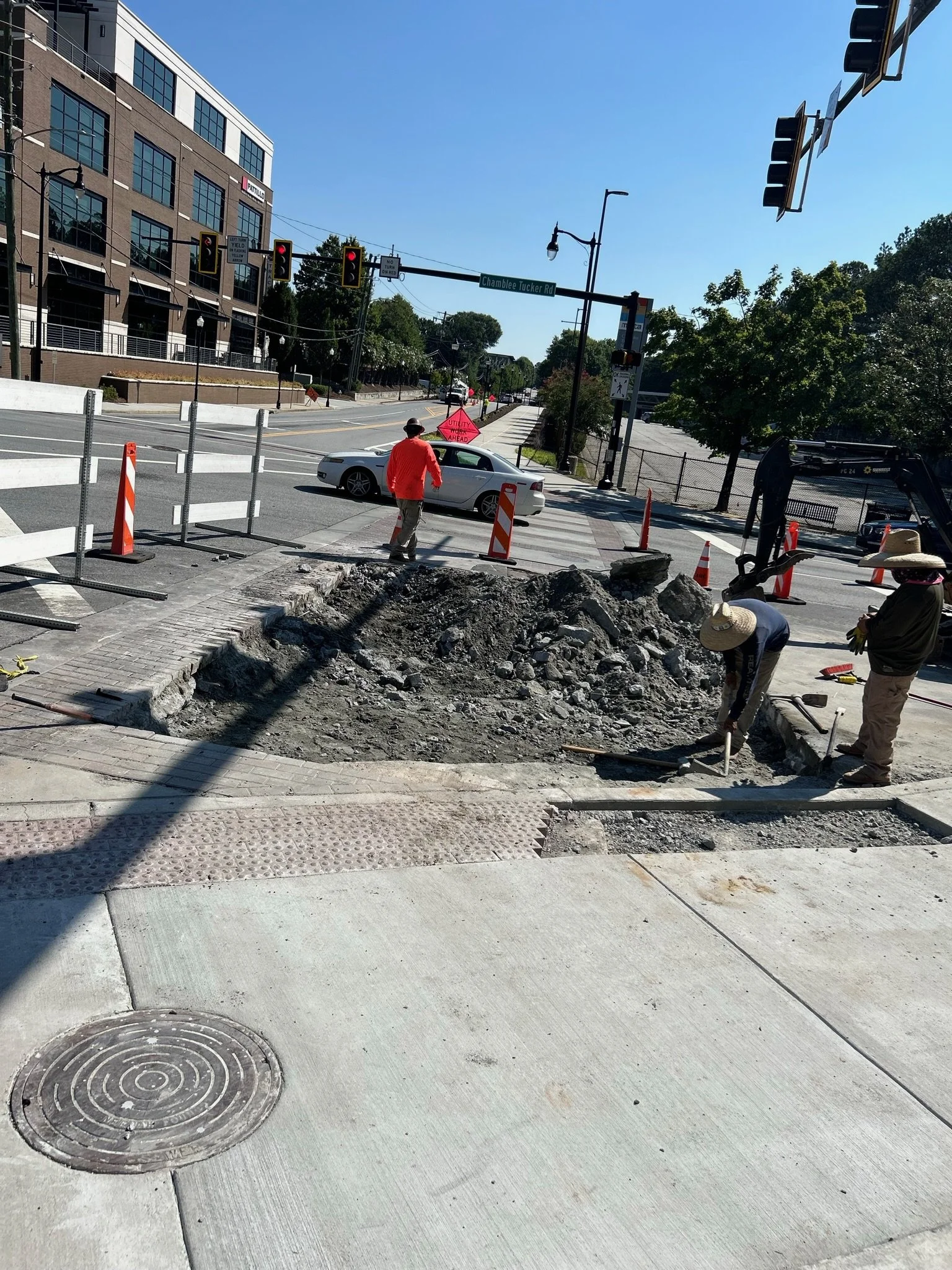 Construction workers repairing a sidewalk with a large hole, surrounded by orange and white traffic cones. A man in a red shirt and hat is walking nearby, while others are working.
