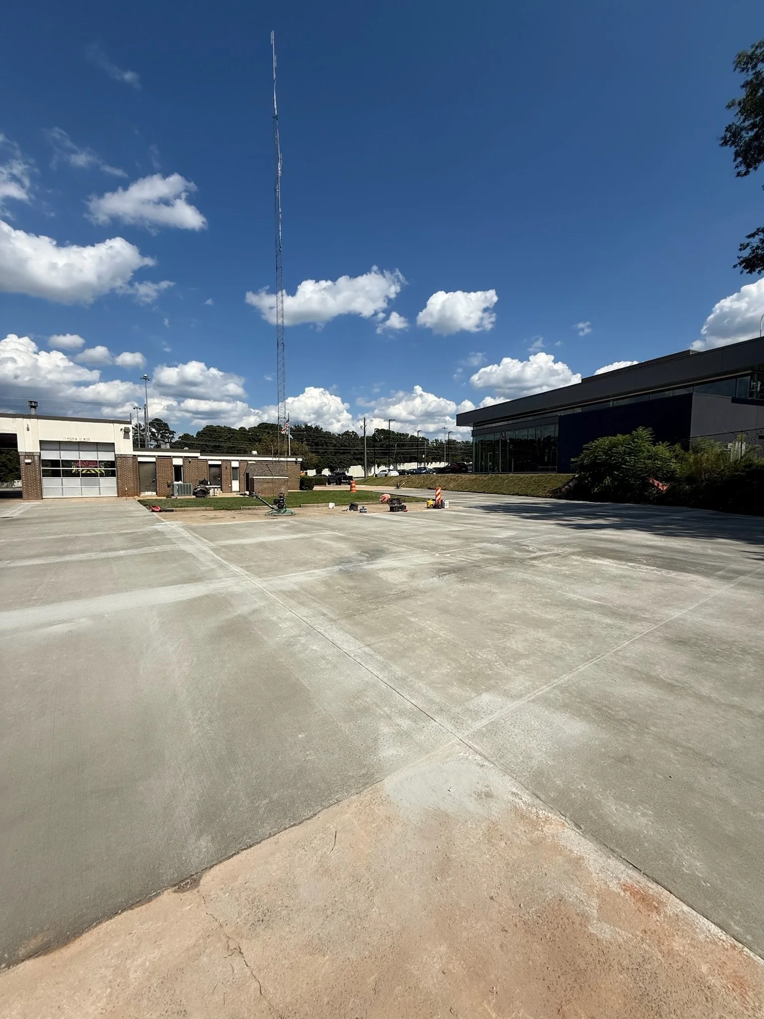 A large, mostly empty parking lot with a building to the right and a smaller brick building to the left. The sky is blue with scattered clouds, and a tall radio tower is visible in the background.