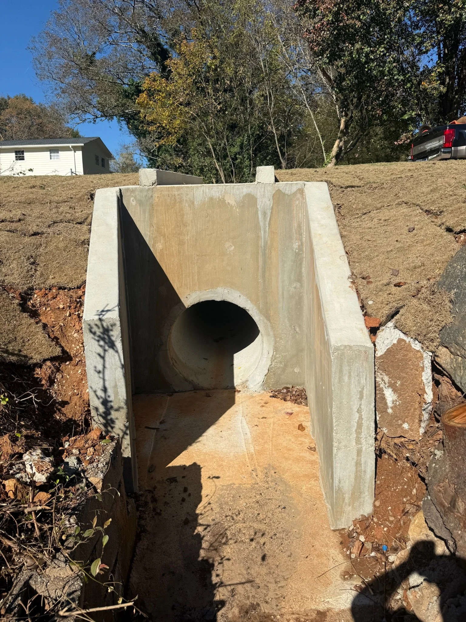 A concrete drainage pipe installed underground in a dirt trench, with a hillside and trees in the background, and a house and pickup truck on the hill.