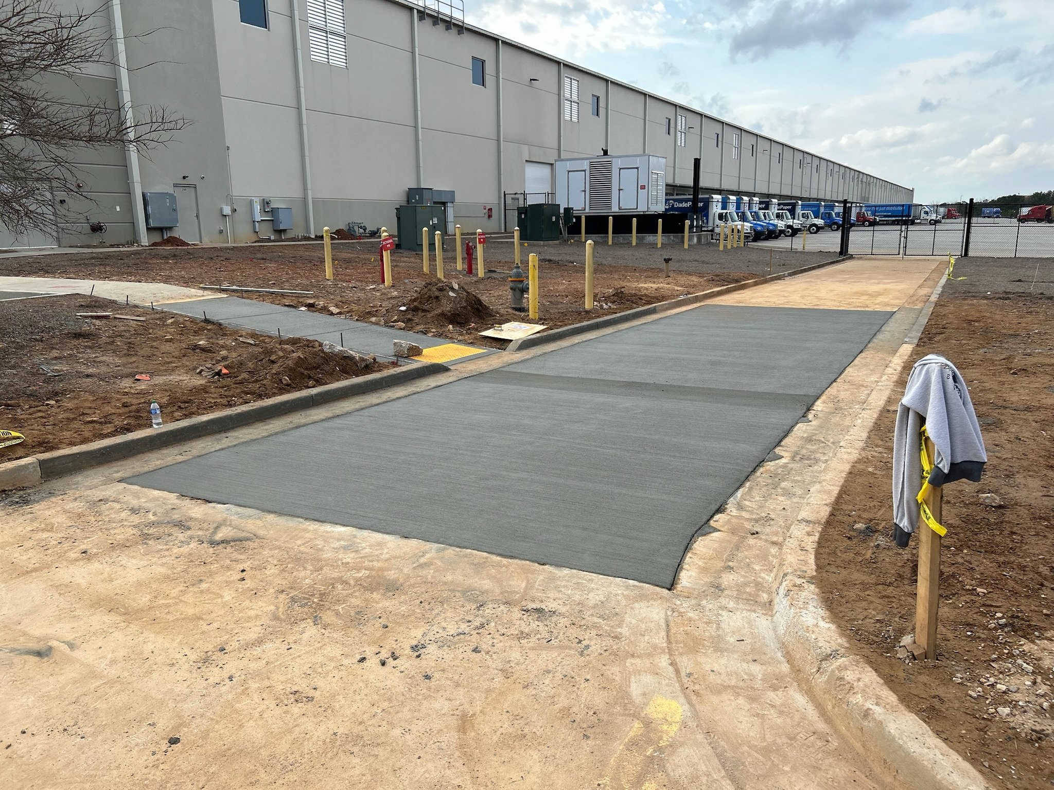 Construction site with freshly poured concrete sidewalks, surrounding dirt, yellow posts, and parked trucks near a large industrial building.