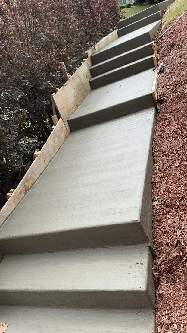 Newly poured concrete stairs with wooden forms on one side, surrounded by red mulch and trees in the background.