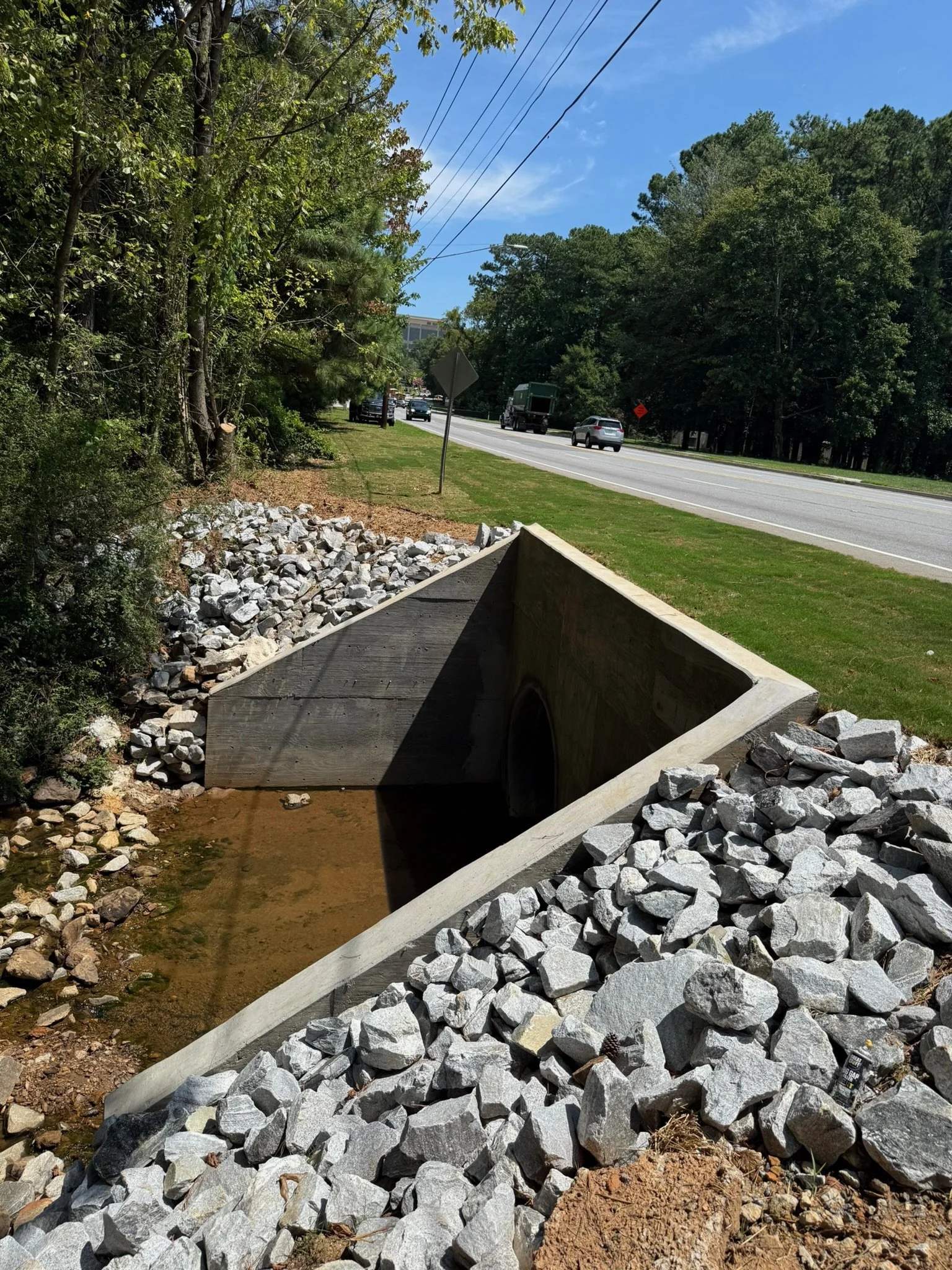 Construction site with large rocks and a concrete drainage ditch beside a grassy roadside and a street with several cars, trees, power lines, and a partly cloudy sky in the background.