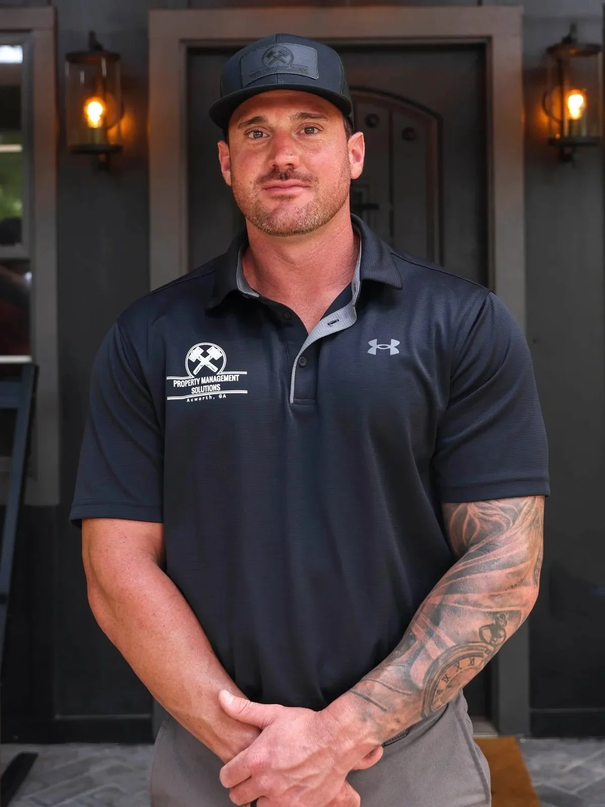 A man in a black cap and a black polo shirt with a company logo, standing indoors near a doorway with warm lighting.